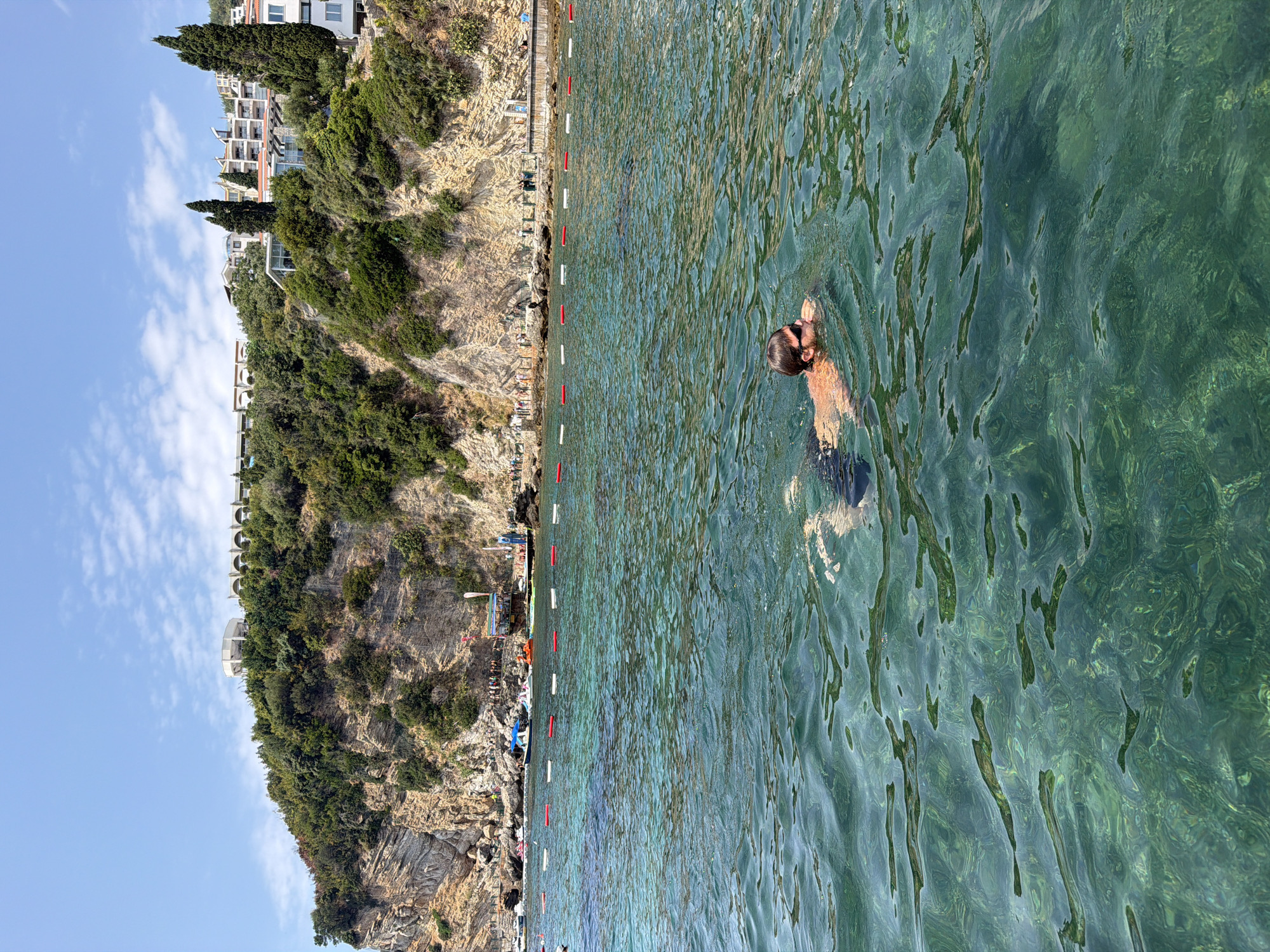 A man swims in the clear, green waters of the Adriatic Sea in Budva, Montenegro. The water is calm and transparent, revealing the rocky seabed below. The man is wearing dark swim trunks and sunglasses. In the background, a steep, rocky cliff rises from the sea, covered in lush green vegetation. On top of the cliff, several white buildings with red roofs are visible, some of which appear to be hotels or resorts. A line of red and white buoys marks the boundary of a swimming area. Several small boats and jet skis are moored near the shore. The sky is a clear blue with a few wispy clouds.