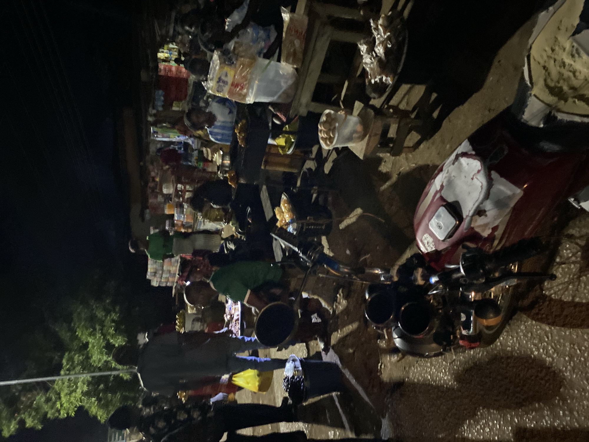 The image depicts a street scene in Bukuru, Nigeria at night. A motorcycle is parked on the right side of the image. Several people are gathered around what appears to be a roadside market stall. The stall displays various items for sale, including packaged goods, fruits, and other food items. Some individuals are interacting with the vendors, possibly purchasing goods. The environment is dark with limited artificial lighting.