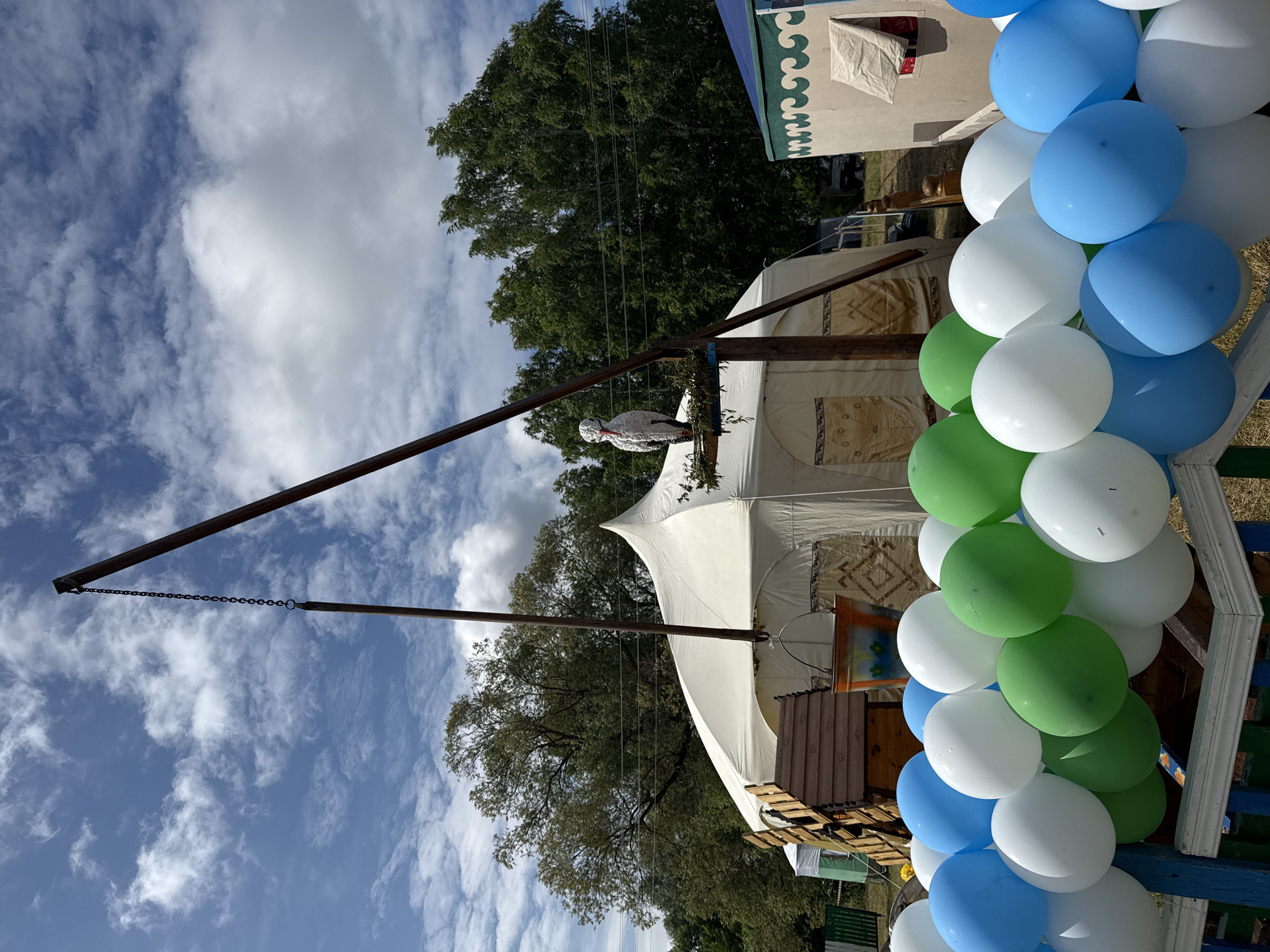In the village of Staroye Subkhankulovo, Russia, a festive outdoor scene unfolds under a partly cloudy sky. Dominating the center is a large, white yurt-like tent, its peak reaching towards the heavens. In front of the tent, a wooden structure resembling a well or a staircase stands, adorned with a decorative bucket hanging from a metal chain. Above this, a large, white, stork-like effigy is perched on a wooden platform, its beak a vibrant red.
The scene is further enhanced by a colorful array of balloons in shades of blue, white, and green, arranged in clusters and forming a vibrant border in the foreground. These balloons are attached to a blue and white fence. In the background, lush green trees create a natural backdrop, with the faint outlines of other structures suggesting a gathering or event. The overall impression is one of a joyous celebration in a rural setting.