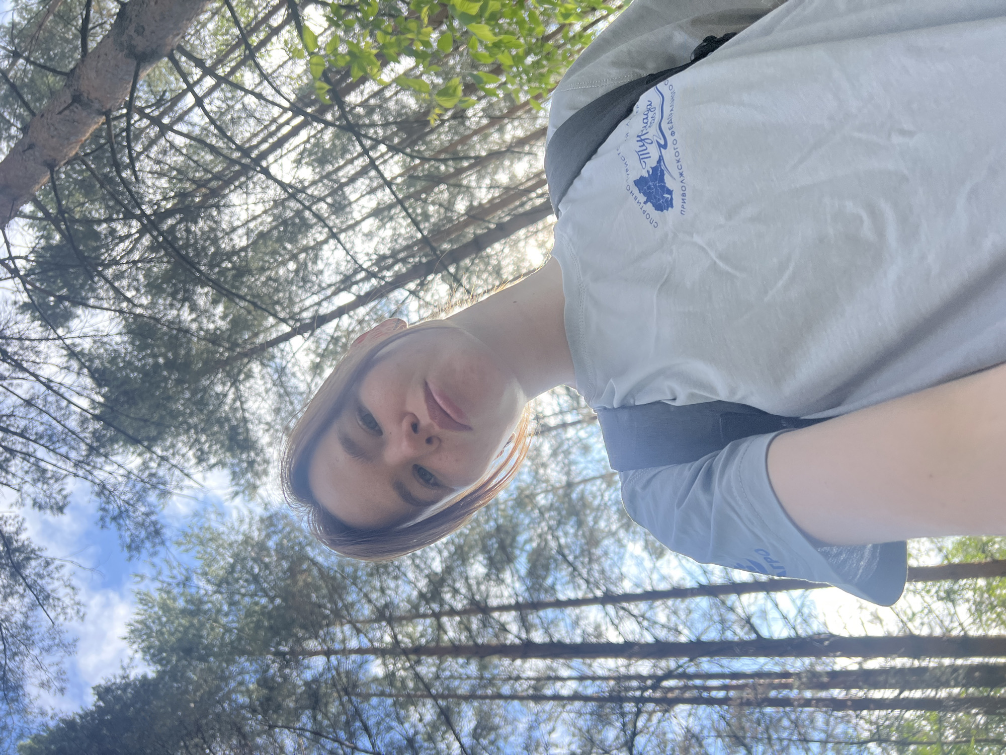 In Chaikovsky, Russia, a young woman with light brown hair is captured in a low-angle, selfie-style photograph taken outdoors amidst a forest. She is wearing a light grey t-shirt with a blue logo featuring text that appears to be in Cyrillic script, and a dark grey backpack strap is visible over her shoulder. The background is dominated by tall, slender pine trees with a bright, dappled sky peeking through the dense canopy, indicating it is daytime with partial sunlight. The perspective looks upwards, emphasizing the height of the trees and the open sky.