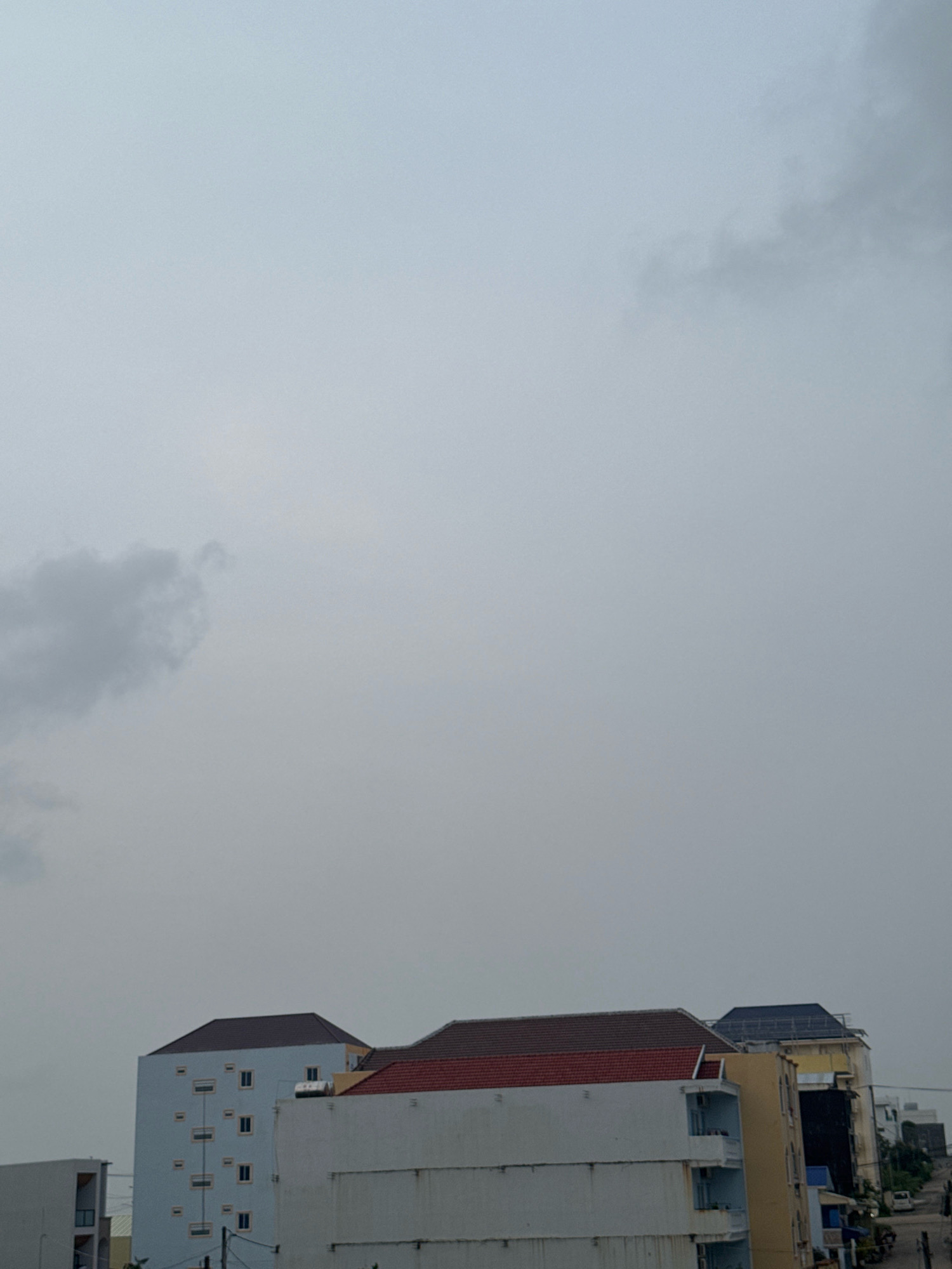 The image shows a cloudy sky over a row of multi-story buildings in Sihanoukville, Cambodia. The buildings are of varying heights and architectural styles, with some featuring balconies and pitched roofs. The sky is predominantly a uniform gray, with some darker clouds visible on the left side of the frame. The overall impression is a somewhat overcast day in an urban coastal environment.