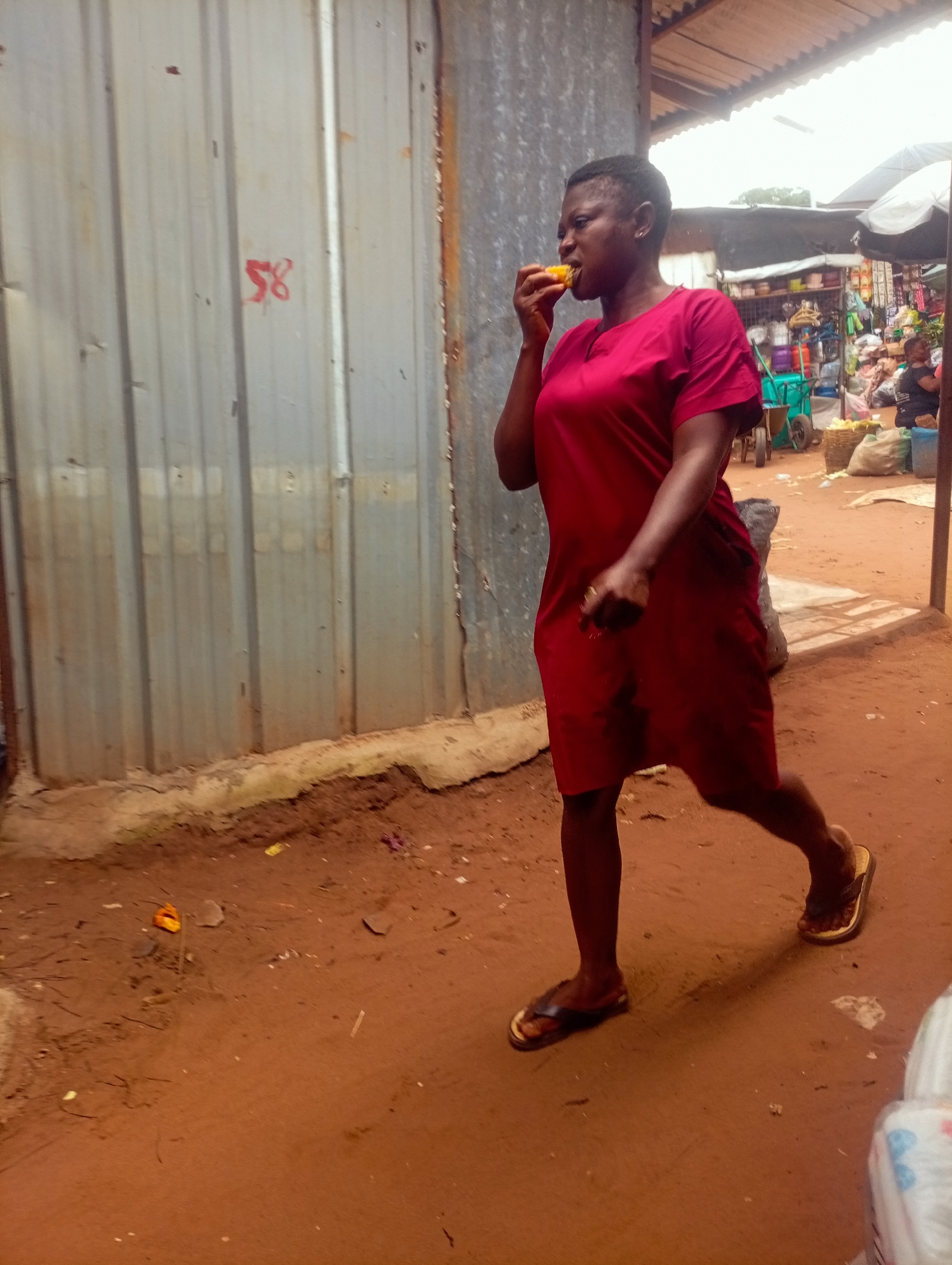 A young woman in a red dress walks past a corrugated metal wall, eating a piece of corn. She has short hair and is wearing flip-flops. The wall has the number "58" spray-painted in red on it. Behind her, a bustling market scene unfolds with stalls, produce, and other people. The ground is made of reddish-brown dirt. The lighting suggests it is daytime.