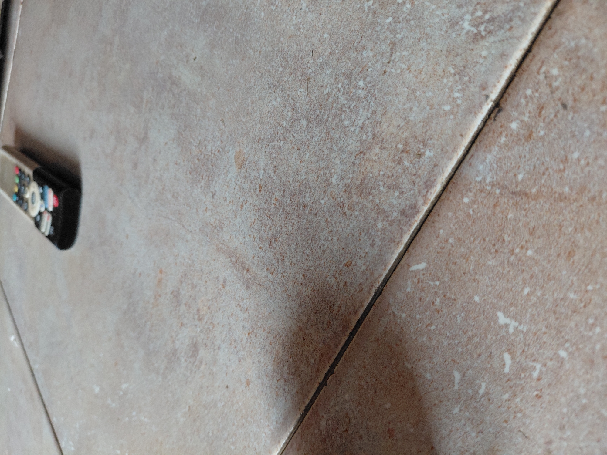 The image presents an indoor scene primarily composed of light brown or beige floor tiles, possibly in Santa Ana, Venezuela. The perspective is low and angled, looking across the tiled surface.
The main subjects are the ceramic or porcelain tiles, which exhibit a speckled, slightly textured appearance with varying shades of tan, brown, and off-white. A prominent dark grout line runs diagonally from the upper left to the lower right of the frame, dividing the tiled surface and creating a strong compositional element. This grout line appears dark brown or black and shows some minor irregularities, suggesting wear or imperfect application.
On the far left side of the image, partially obscured and out of focus, lies a remote control. It is black and silver with a variety of small, colorful buttons, including white, blue, green, red, yellow, and black. Due to the blur and angle, no discernible text or specific symbols on the remote's buttons are visible.
There are no people present, and no specific activity or event is taking place; the scene depicts static objects. The lighting appears to be ambient indoor light, and there are no cues to determine the time of day or specific weather conditions. The overall impression is one of an everyday domestic or interior space.