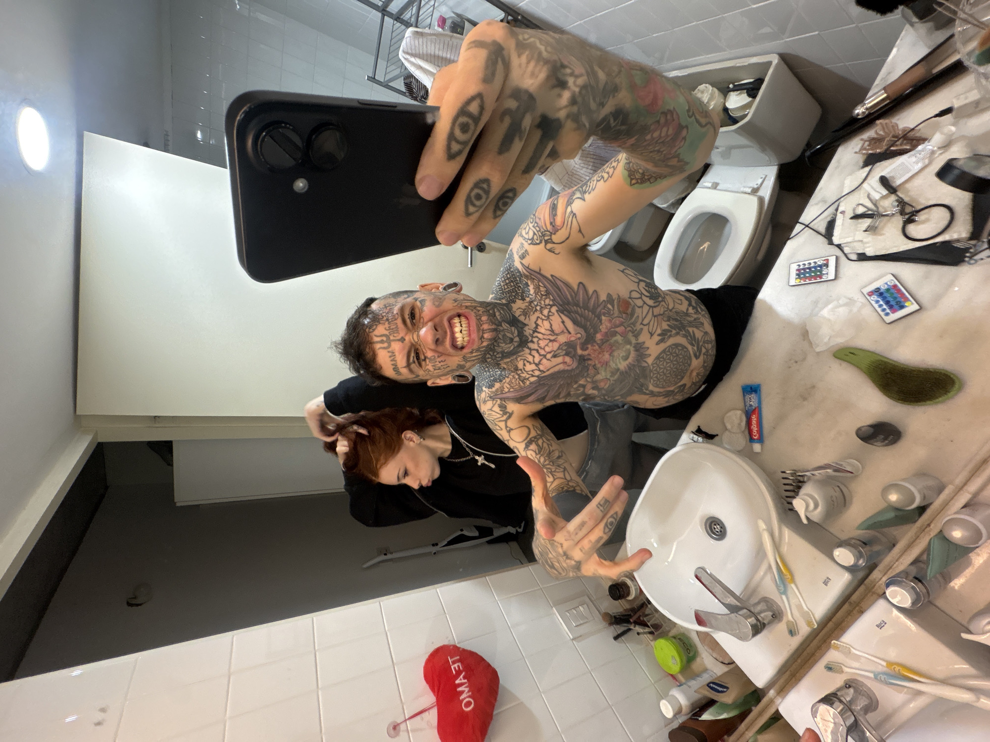 The scene takes place in a residential bathroom in Buenos Aires, Argentina. A wide-angle mirror selfie captures two individuals.
In the foreground, a male subject, heavily tattooed on his face, neck, chest, and arms, holds a black smartphone to take the photo. He has dark, short hair, prominent ear gauges, and a wide, intense grin revealing his teeth. His chest features a large eagle tattoo, and his left hand, extended towards the camera with fingers spread, displays multiple eye tattoos on the knuckles and fingers. He appears shirtless from the waist up.
Behind and slightly to the left of the male, a female subject with reddish-brown hair is visible. She uses both hands to gather her hair, with a slight pout on her face. She wears a black long-sleeved top and a silver cross pendant.
The bathroom features white tiled walls and a white ceramic sink with a single-lever faucet. The countertop around the sink is cluttered with various toiletries, including bottles, tubes, two toothbrushes (one yellow, one blue), toothpaste, and a green hairbrush. A red plush heart with the text "OMAЭТ" (likely a stylized "TE AMO") hangs on the tiled wall to the left.
Behind the male subject, a white toilet with its lid open and tank cover removed is visible, exposing internal mechanisms. The countertop to the right of the sink holds two small rectangular remote controls with colorful buttons, hair tools, scissors, and other miscellaneous items. The reflection in the mirror reveals a metal shelving unit with towels, a vacuum cleaner, and a dimly lit adjacent space with a white appliance. Overhead recessed lighting is also visible in the reflection.