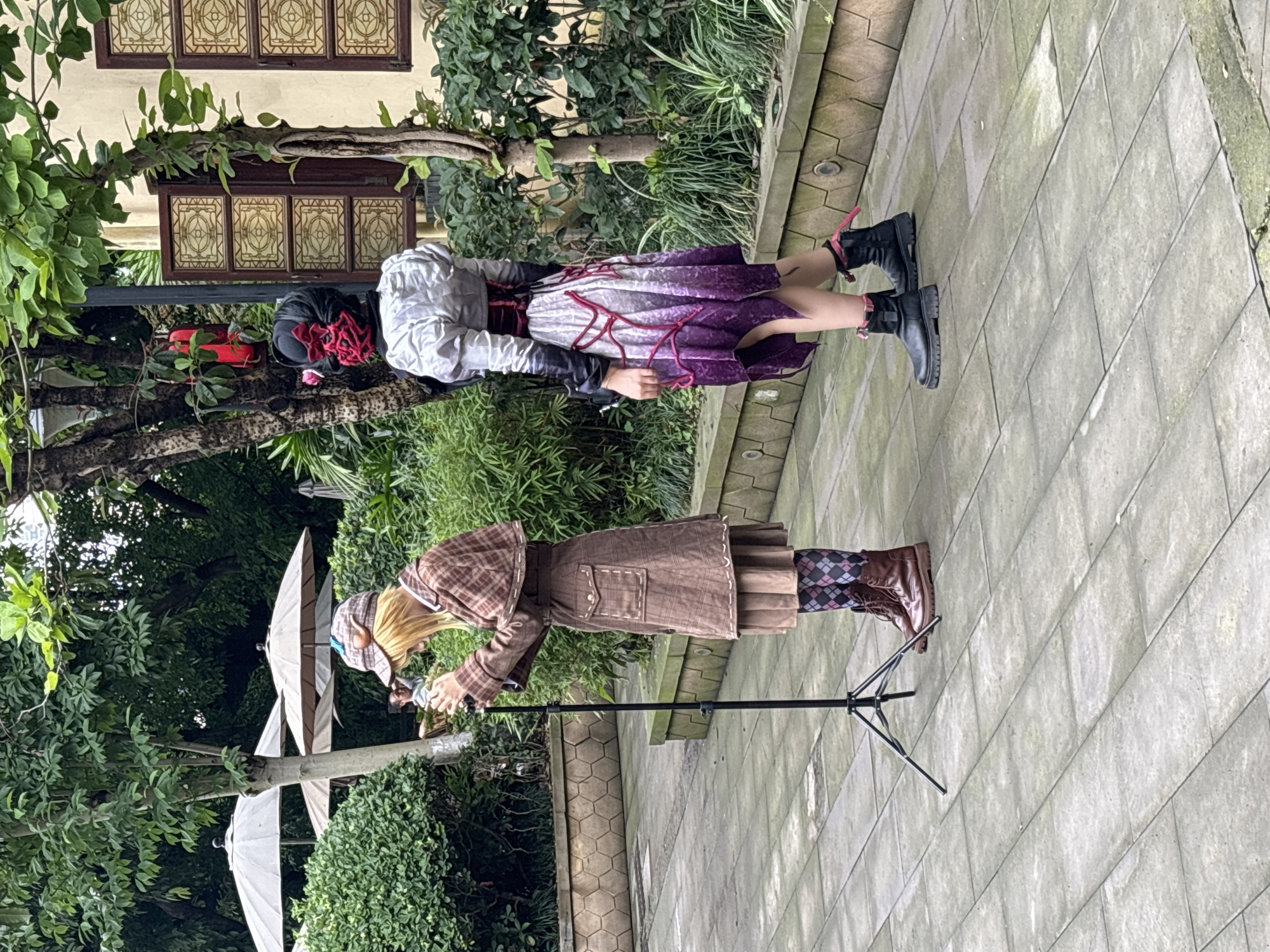 Two people are present in a public outdoor space. One individual is attired in a long brown coat, a hat and brown boots with patterned tights. The other is in a dress with purple and white hues, a white blouse, and black boots. A microphone stand is also in view. The location is in Yuzhong District, Chongqing, China.