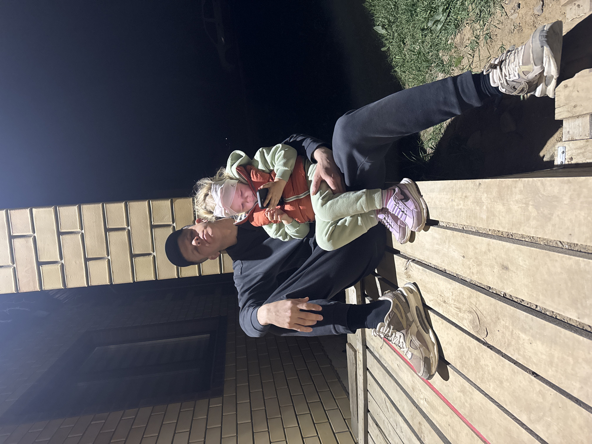 A man is sitting on a wooden surface, holding a young child. The man is wearing black clothing, including a cap. The child is bundled in a light green hooded sweatshirt and an orange vest, with a white head covering. They are holding a smartphone. The scene is at night, next to a brick building. The location is Ферма, Russia.