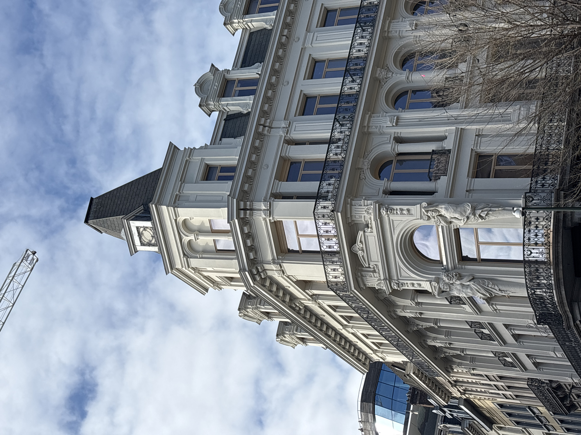 The image shows a tall, ornate, light-colored building in Brussels, Belgium, viewed from a low angle, tilting upwards. The building features multiple stories with a variety of window shapes (rectangular and arched), decorative moldings, balconies, and sculptural elements. A modern, blue-glass building is visible at the bottom of the frame, suggesting a contrast between old and new architecture. A crane is visible in the sky. The sky is cloudy with patches of blue. Bare tree branches appear on the right side of the frame. No people are visible.