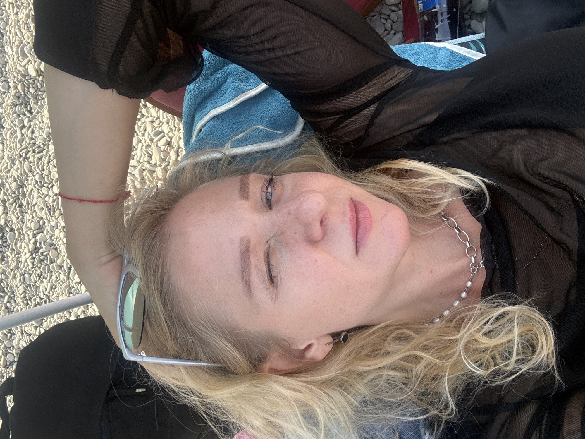 A blonde woman is lying on a pebble beach in Partenit, Ukraine. She is wearing a sheer black top and a silver necklace with pearls. She is looking at the camera and winking. Sunglasses are resting on her head. A blue towel is visible behind her.