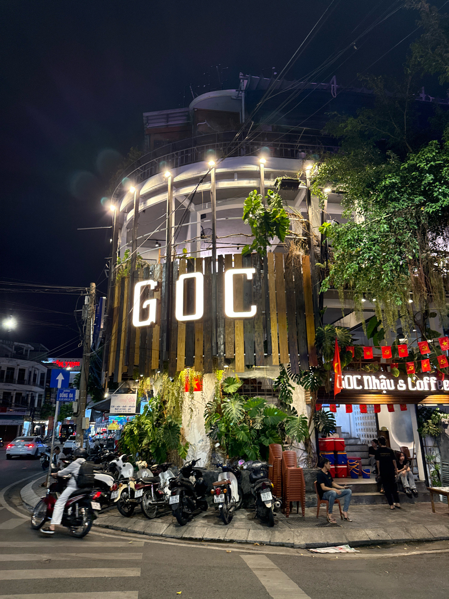 The image depicts a night scene in front of a coffee shop named "GOC Nhậu & Coffee" located in Nha Trang, Vietnam. The coffee shop features a prominent illuminated sign with the word "GOC" in large white letters. The building has a multi-level structure with outdoor seating and is adorned with plants and hanging foliage. Red decorative flags with yellow stars are strung across the facade. Several motorcycles are parked on the sidewalk and street in front of the establishment. A few people are visible, including patrons sitting outside and staff members. In the foreground, a pedestrian crossing is marked on the street, and a person is riding a motorcycle. The street is illuminated by artificial lights, and the sky is dark, indicating it is nighttime.