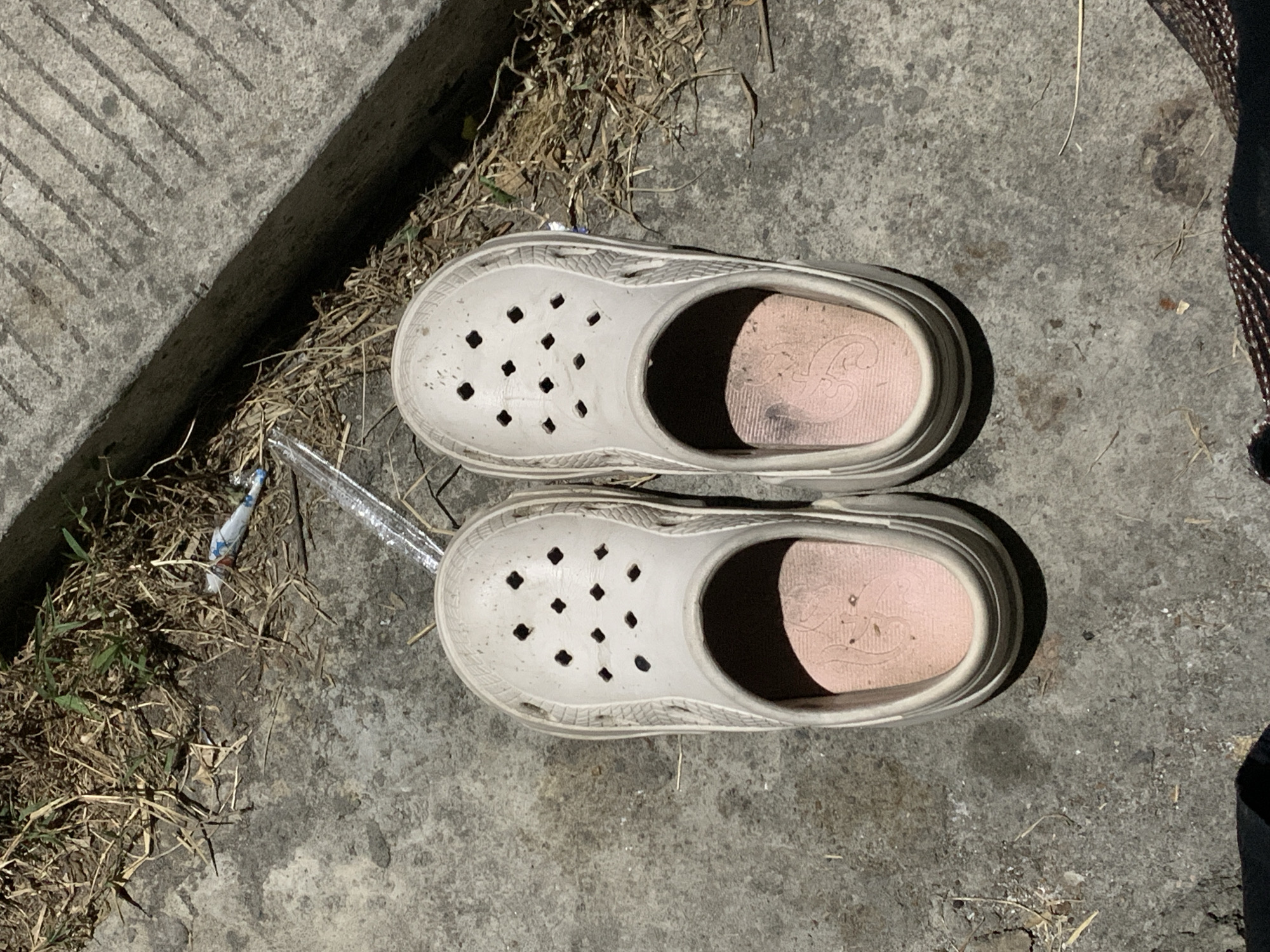 In Jungke, Indonesia, a pair of worn, light beige Crocs-style clogs with pink insoles featuring an embossed design rests on a mottled grey concrete surface. The shoes are oriented with their toes pointing towards the upper frame. The concrete is stained and sparsely covered with dry organic debris and small fragments of trash, including a crumpled colored wrapper (blue, white, red) and a clear plastic shard. To the upper left, a patch of dried grass with some green sprouts is visible. Along the top and right side of the frame, a concrete curb with parallel grooves casts a distinct shadow. The scene is illuminated by strong, direct daylight, creating prominent shadows. No individuals are present.