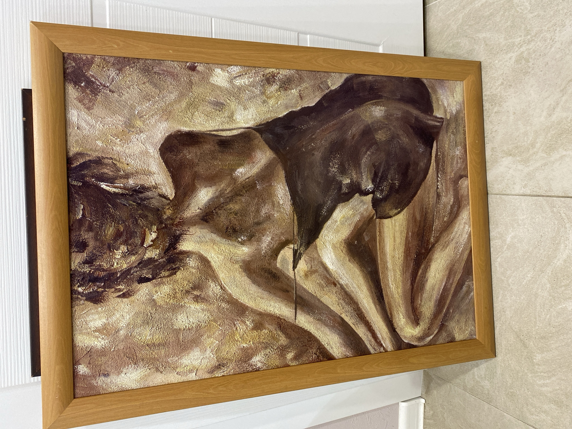 A framed oil painting is mounted on a white door next to a tiled wall, appearing to be indoors. The painting features figures rendered in browns, tans, and yellows with broad brushstrokes, suggesting a draped or reclining subject. The frame is a simple, light wood color.