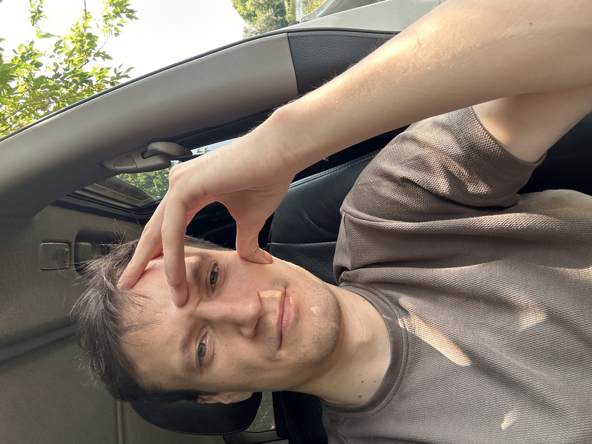 A man with brown hair and light skin is sitting in the driver's seat of a car. He is wearing a brown t-shirt. He has his right hand on his forehead, with his fingers extended. His left arm is resting on the car door. The car appears to be parked outdoors, with trees and a body of water visible in the background. The sun is shining, casting shadows on the man's face and shirt. The image was taken in Makiv, Ukraine.