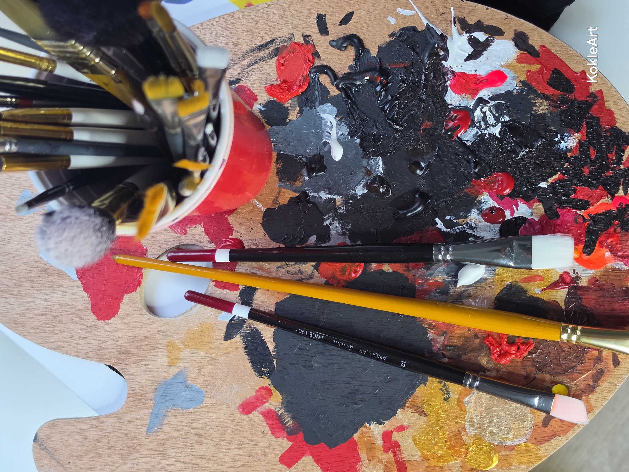 A top-down, close-up view showcases an artist's wooden palette covered in various splotches and smears of paint in red, black, white, and yellow. Three paintbrushes are positioned on the palette: one black-handled brush with a pink tip, a yellow-handled brush, and another black-handled brush with a white tip. A red cup filled with numerous paintbrushes is placed at the top right of the palette. The environment is an indoor setting, likely an art studio, but its specific location in Melhus, Norway, cannot be determined from the image alone. There are no people or interactions depicted in the scene.