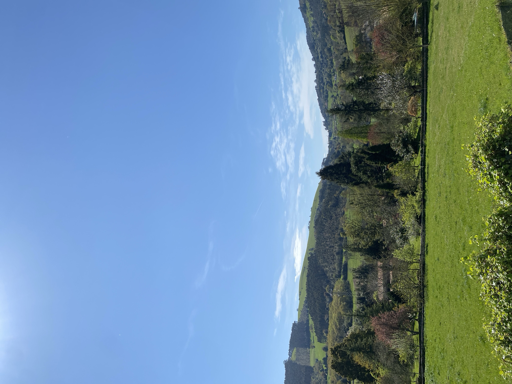 The image shows a landscape with green hills, trees, and a clear blue sky. The scene is located in Villaviciosa, Spain, as evidenced by the architectural style and vegetation common to the region. The foreground features a lush green meadow bordered by a fence line and various shrubs and trees. Beyond the meadow, the hills rise with mixed forest cover, including deciduous and coniferous trees. The sky is predominantly blue with scattered clouds. The sun is positioned on the left side of the image, creating a subtle glare. The overall impression is of a serene, rural setting.