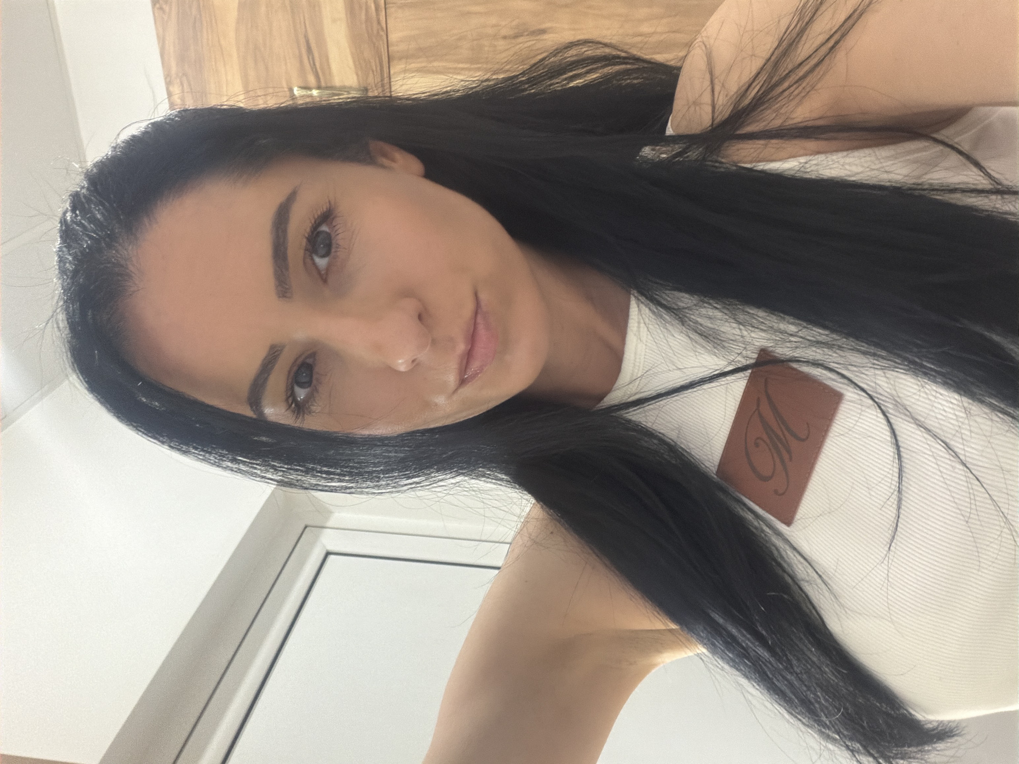 An adult female is depicted in a medium close-up, facing the camera directly, in what appears to be an indoor setting. She possesses long, dark hair parted slightly to her left, dark eyes, and a fair complexion, with a neutral facial expression and slightly glossy lips. The subject is wearing a light-colored, ribbed sleeveless top featuring a rectangular brown patch with a black, stylized "M" located on the upper left chest area. Her left arm is partially visible, raised. The background consists of a white door and frame to the left of the subject, and a wooden panel or door to her right. The lighting is bright and artificial. The precise location within the city of Pleven, Bulgaria, cannot be determined from the visual information provided.