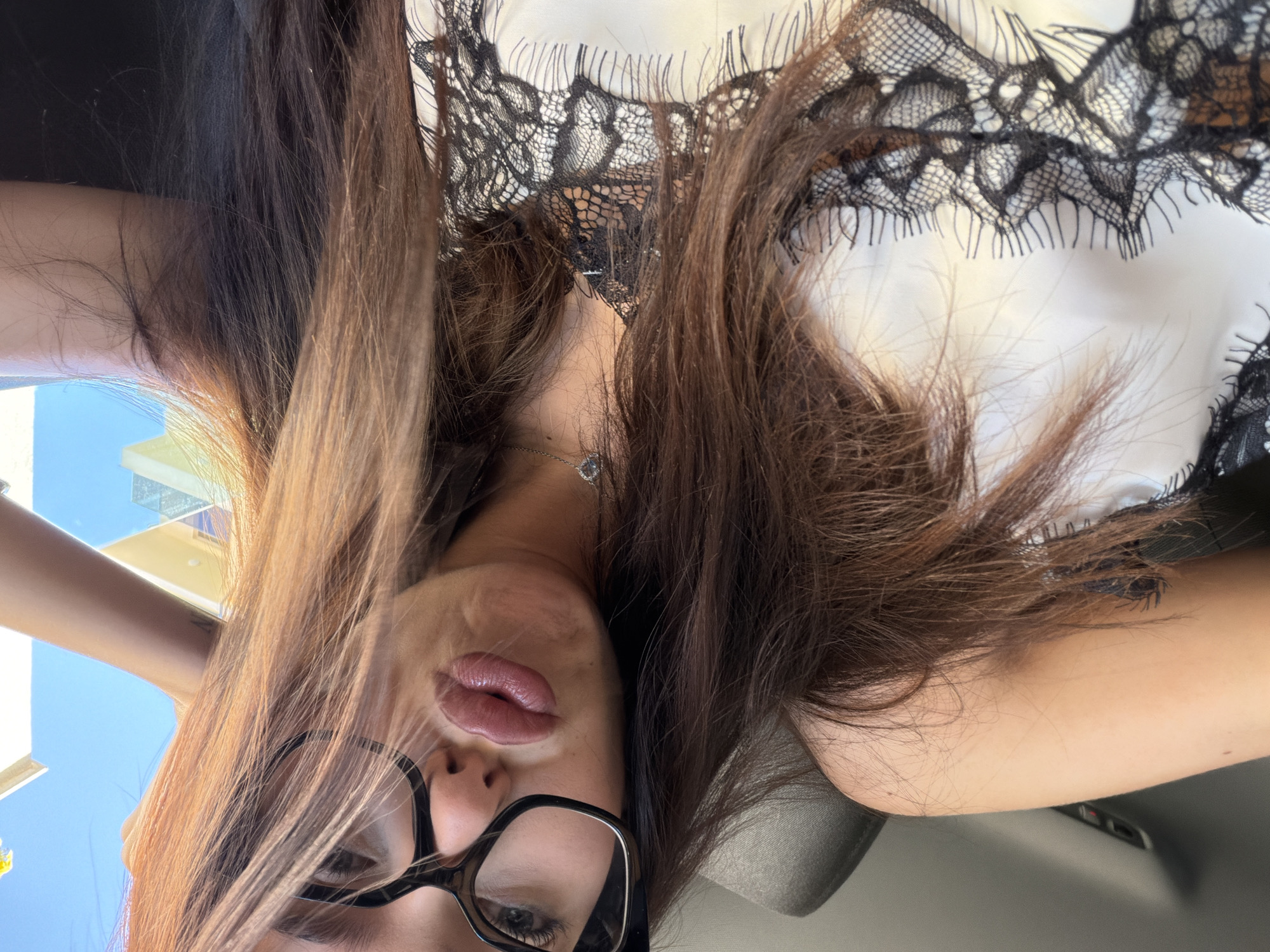 A woman is taking a selfie while sitting in a car. She is wearing a white camisole with black lace trim and large black-rimmed glasses. Her lips are puckered, and her hand is resting on her head. Her hair is long and brown, with some strands falling over her face. The background shows a blue sky and part of a building.