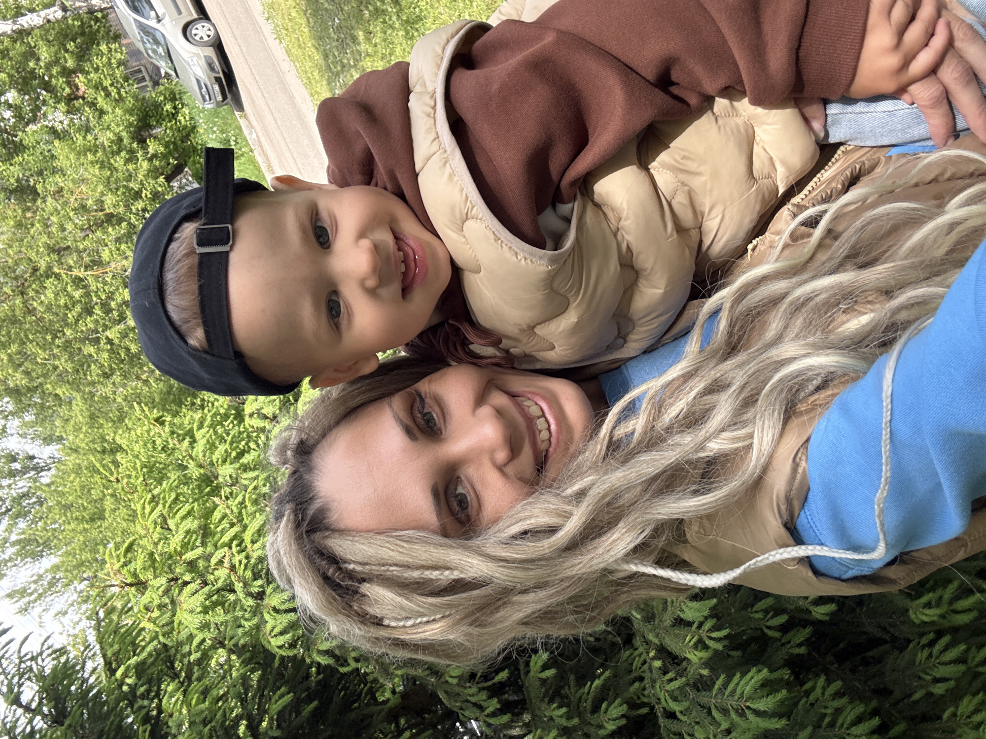 A woman and a young child are in the foreground. The woman has blonde hair with long braids. She is wearing a light blue sweater and a tan vest. The child, who has a black baseball cap, is wearing a brown sweatshirt and a tan vest. They are both smiling at the camera. A stand of trees is on the left side of the image, and a silver car is parked at the edge of a sidewalk. The photo was apparently taken while lying down. It is impossible to confirm the location is Rusanovo, Russia, from the image.
