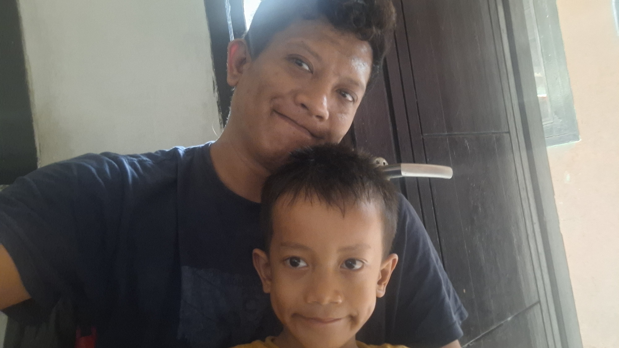 A selfie is taken of a man and a child. The man is in the background, looking at the camera with a slight smile. He is wearing a dark blue t-shirt. The child is in the foreground, also looking at the camera with a smile. The child is wearing a yellow shirt. The background consists of a dark wooden door and a light-colored wall. The image appears to be taken indoors, possibly in a residential setting in Cibinong Hilir, Indonesia.