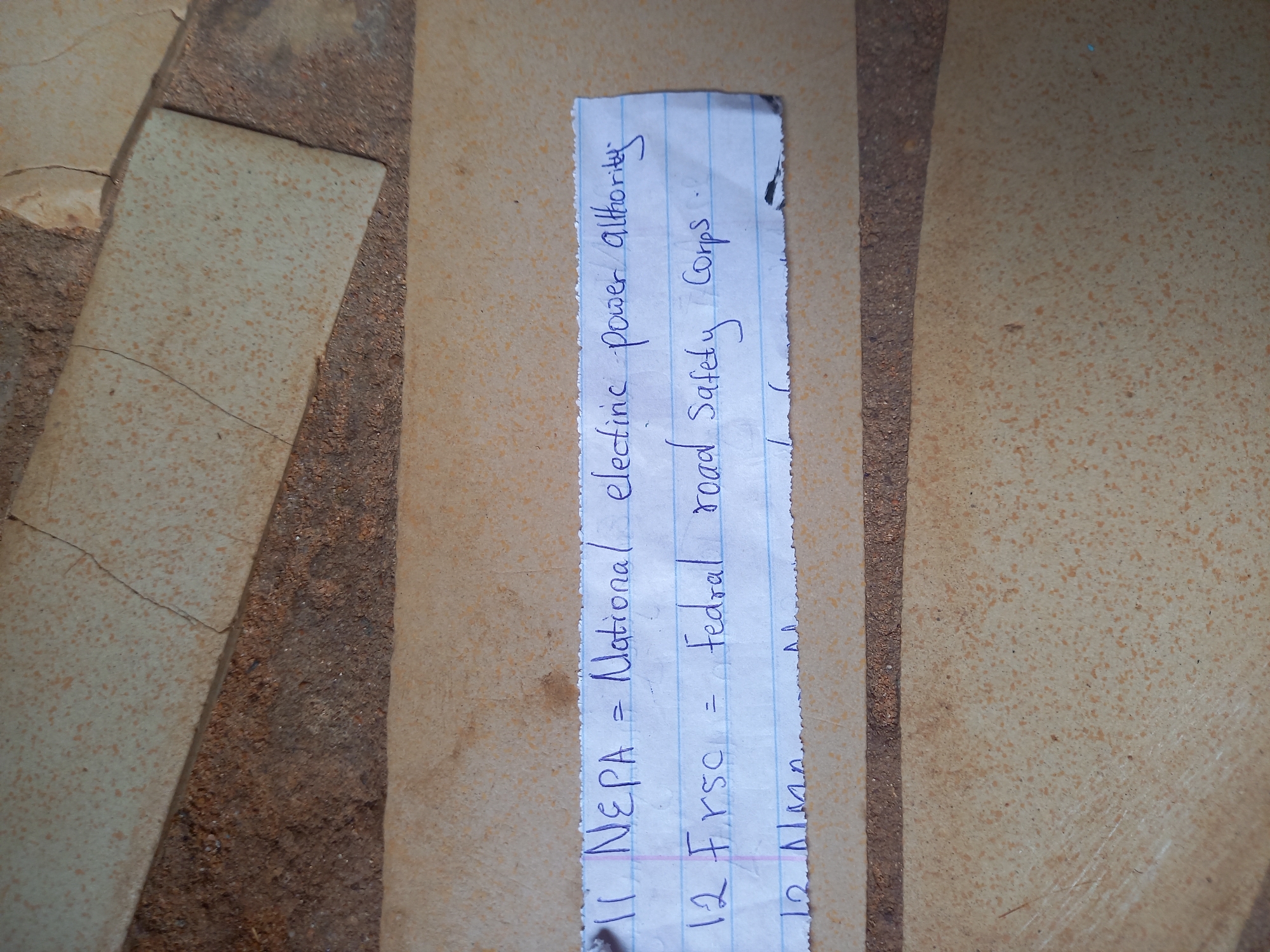 The image captures a close-up view of a small, irregularly torn piece of lined white paper resting on a textured ground surface. The paper contains handwritten text in blue ink, listing acronyms and their full forms. The ground itself is composed of light brown and beige-mottled concrete or tiled surfaces, interspersed with darker, granular material filling cracks and gaps, suggesting an outdoor or semi-outdoor setting.
The visible text on the paper is written horizontally and includes:
- "11 NEPA = National electric power authority" (with "electric" and "authority" slightly misspelled in the original writing as "electinic" and "althority" respectively).
- "12 FRSC = Federal road Safety Corps" (with "Federal" slightly misspelled in the original writing as "Fedral").
- A partial entry numbered "13" followed by an acronym beginning with "AA" is visible at the bottom edge of the paper, but its full content is cut off.
The lighting appears bright and even, indicative of daytime. There are no people visible, and no specific activity is taking place other than the paper lying on the ground. The paper's torn edges suggest it might have been ripped from a larger document or discarded. The setting, identified as Onitsha, Nigeria, aligns with the acronyms which are relevant to Nigerian organizations (NEPA - National Electric Power Authority, FRSC - Federal Road Safety Corps). The overall impression is that of a casual, possibly discarded, note lying on a typical paved surface.