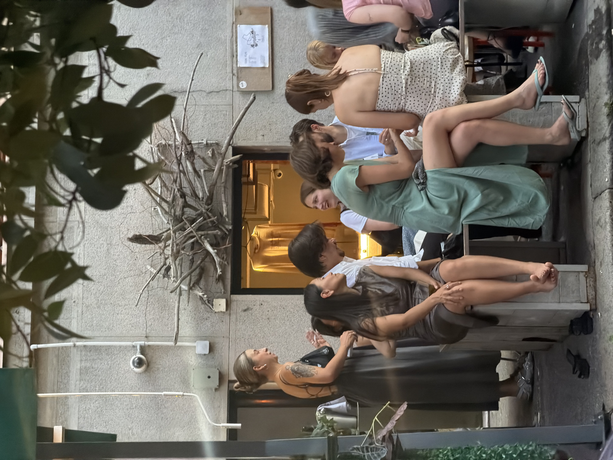 In Belgrade, Serbia, on a wall with a light grey textured finish, a CCTV camera is mounted. Below it, a green electrical box is attached. A wooden structure made of branches is positioned above a doorway that leads into a space where a yellow garment is displayed on a hanger. In front of this wall, a group of people are gathered. A woman in a dark grey dress, with her back to the camera, is standing and holding a purse. To her right, a woman with dark hair is seated. In the center, a man wearing a white shirt is talking to a woman in a light green dress. To the right of them, a woman with red hair in a polka dot dress is seated and facing away from the camera. The people are sitting on or near wooden benches and seating. The scene appears to be an outdoor seating area, possibly a cafe or restaurant.