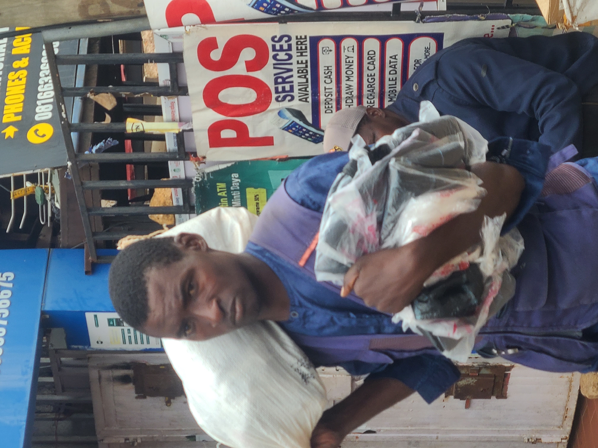 In the city of Jos, Nigeria, a man of African descent, appearing to be in his late 30s or early 40s, walks carrying two large loads. The primary load, a large white sack, is slung over his left shoulder, with its bulk resting on his upper back and left side. His right arm is bent, holding a second load, which consists of multiple items wrapped in clear plastic. These items appear dark, with hints of red and white packaging visible through the plastic. The man is wearing a blue tunic with a V-neck and a purple vest over it. The vest has several pockets and a central zipper.
The background suggests an outdoor urban setting. Behind the man, a fence made of dark metal bars is visible. Beyond the fence, there are signs and structures typical of a commercial area. One prominent sign, partially obscured, features large red lettering spelling "POS". Below this, smaller text reads "SERVICES AVAILABLE HERE" and lists options such as "DEPOSIT CASH", "WITHDRAW MONEY", "RECHARGE CARD", and "MOBILE DATA". To the left of the "POS" sign, another green sign displays text including "tin ATM" and "Minti Daya". Further in the background, a blue building or structure has white text on it, with the numbers "6675" clearly legible at the top. The lighting indicates it is daytime, possibly with overhead shade from structures or the fence. Another person, wearing a dark blue jacket and a cap, is partially visible behind the main subject and to the right. The ground appears to be paved with reddish-brown tiles.