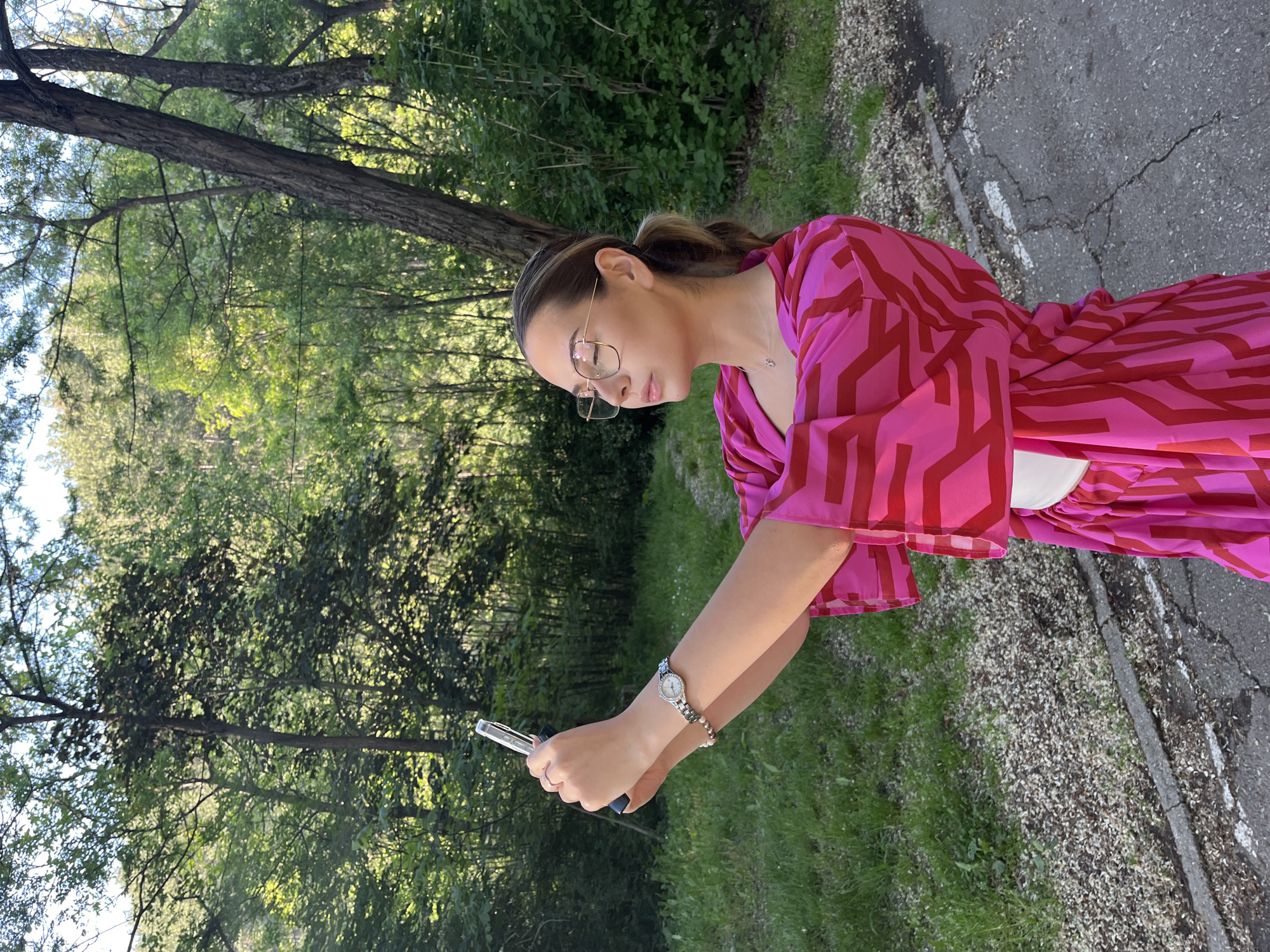 A woman is photographed outdoors in Blagoevgrad, Bulgaria. She is wearing glasses and a pink and red patterned dress with a white waist tie. She is holding a mobile phone in her hands, looking down at it. A wristwatch is visible on her left wrist. The background features a green space with trees, suggesting a park or natural area. To the right of the woman is a cracked concrete pathway.