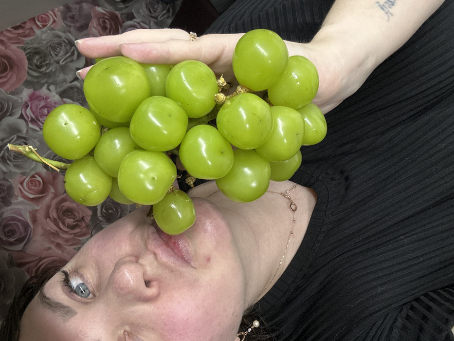 An adult female is partially depicted in an indoor setting. Her face, showing blue eyes and full lips, is angled from the upper left, with her left cheek pressed against one of the green grapes. She wears a dark, ribbed top, a delicate necklace with small ornaments, and a single dangle earring with a pearl-like bead on her left ear. Her right hand, visible in the center-right, supports a large cluster of approximately thirty large, round, glossy green grapes, attached to a visible stem. A ring is present on her right ring finger, and an indistinct tattoo is visible on her inner right forearm. The background consists of a wall covered with dark wallpaper featuring a repeating pattern of stylized pink and red roses. The specific location within the city of Noviy, Russia, is not discernible from the visual content of the image.