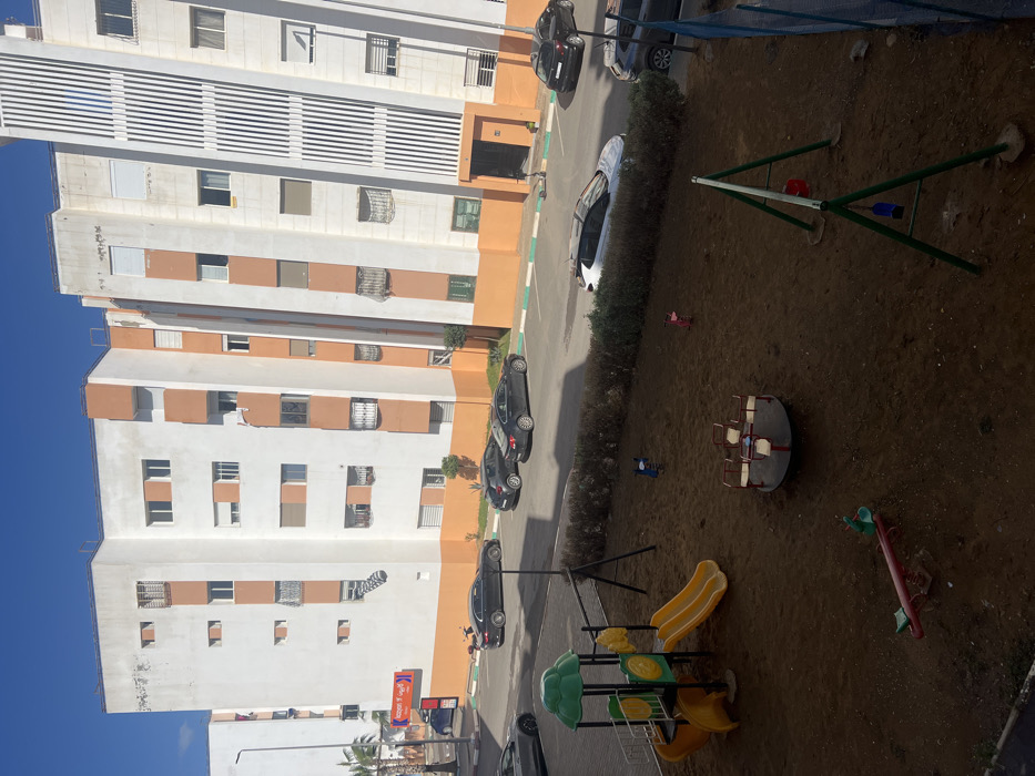 The image depicts an outdoor scene in a residential area of Skhirat, Morocco, under a clear blue sky. In the foreground, a children's playground features brown dirt ground, partially shaded. Equipment includes a green and yellow play structure with two yellow slides, a red and white merry-go-round, a green metal swing set with two empty swings (one red, one blue), a red seesaw, and two empty animal-shaped spring riders (one blue, one pink).
A low hedge separates the playground from a paved street or parking area in the midground. Several vehicles are parked: two dark sedans are parked parallel on the left, a white SUV with a dark roof is parked parallel behind the hedge, and two dark vehicles (one sedan, one SUV) are parked diagonally on the right. Parking spaces are delineated by green and white lines on the asphalt. A solitary, dark-clad individual is visible near the entrance of the rightmost building.
The background is dominated by multi-story residential buildings, primarily white with orange horizontal accent panels around windows and balconies. Many windows feature security bars, decorative grilles, or closed white roller shutters. The building on the far right exhibits distinctive vertical white striped paneling on its facade. On the ground floor of the leftmost building, a commercial establishment with a red awning and "Marjane Express" signage is visible. A street light pole and palm trees are present on the far left. The scene is brightly lit by natural sunlight, with shadows indicating the sun's position.