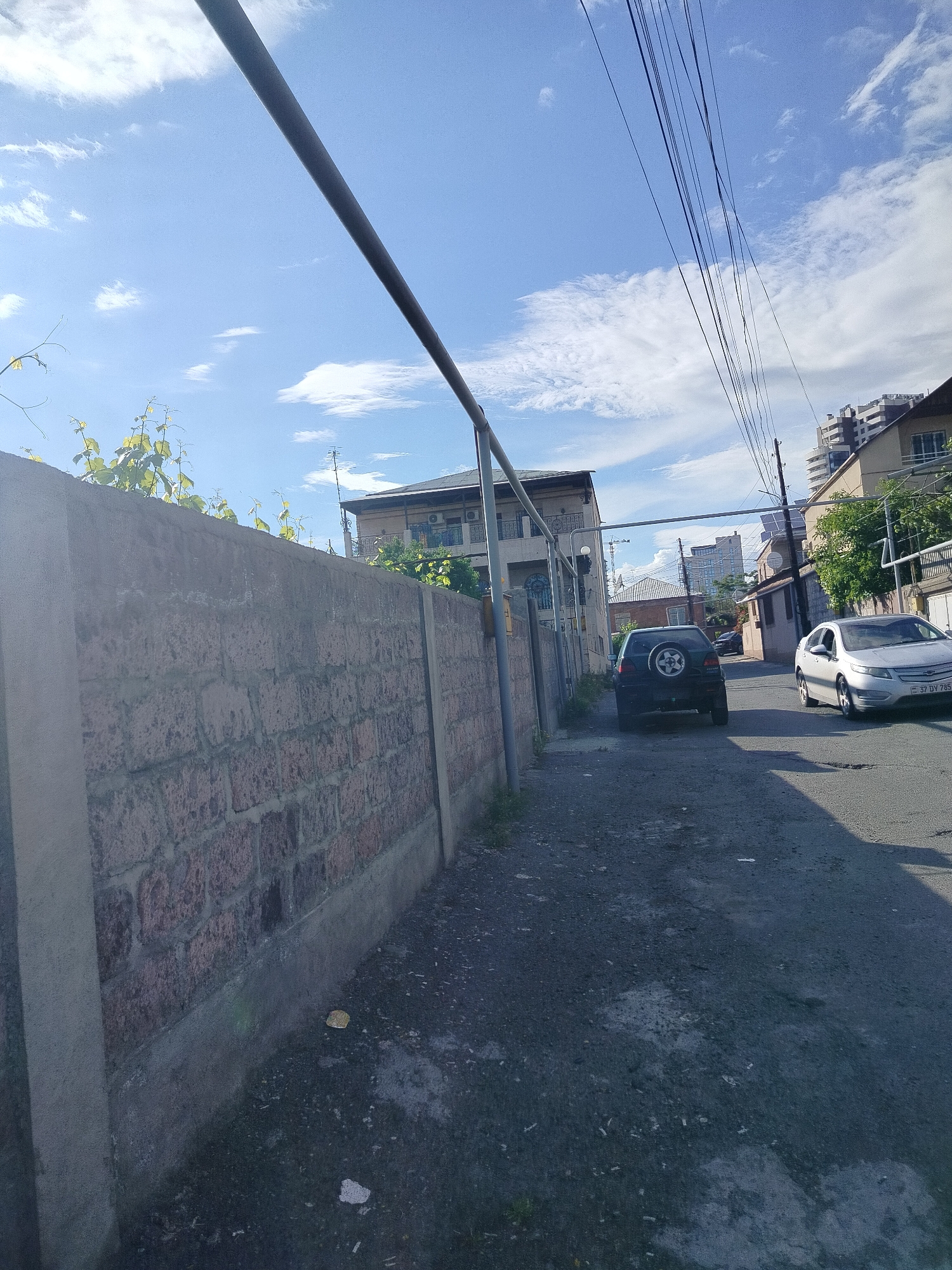 The image depicts a narrow street in Yerevan, Armenia. A brick wall lines the left side of the street, topped with greenery. An overhanging metal pipe runs above the wall. Two cars are visible: a dark-colored SUV with a spare tire mounted on the back and a silver sedan. Power lines stretch across the sky above the street. Buildings of varying heights are visible in the background. The street is paved with a rough, dark surface. The sky is blue with scattered clouds.