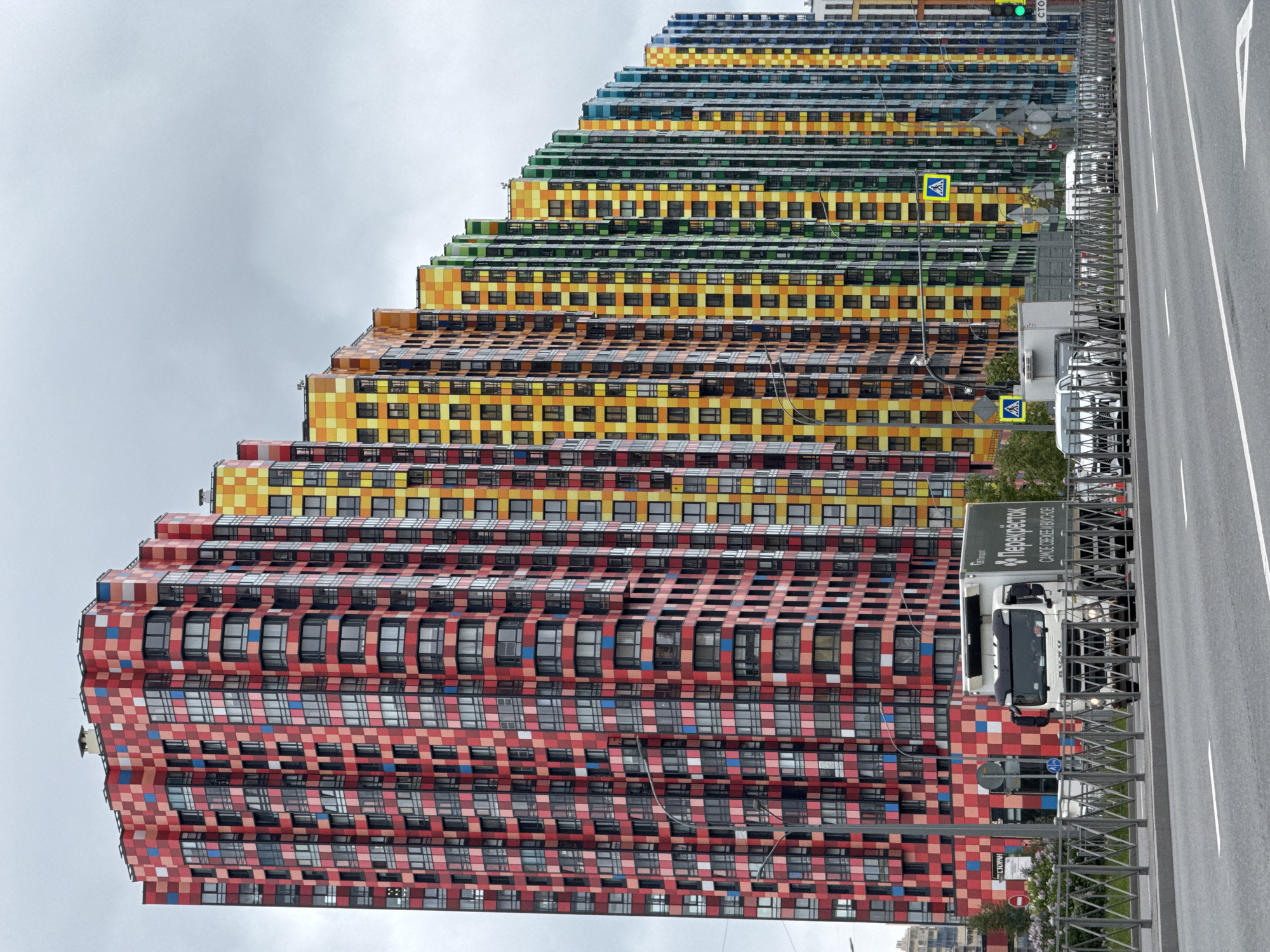 The image shows a row of colorful residential buildings in Saint Petersburg, Russia. The buildings are notable for their terraced design and the use of vibrant, pixelated color schemes ranging from red to blue. The street runs along the side of the buildings and a white truck is visible near a pedestrian crossing sign. The sky is overcast and grey.