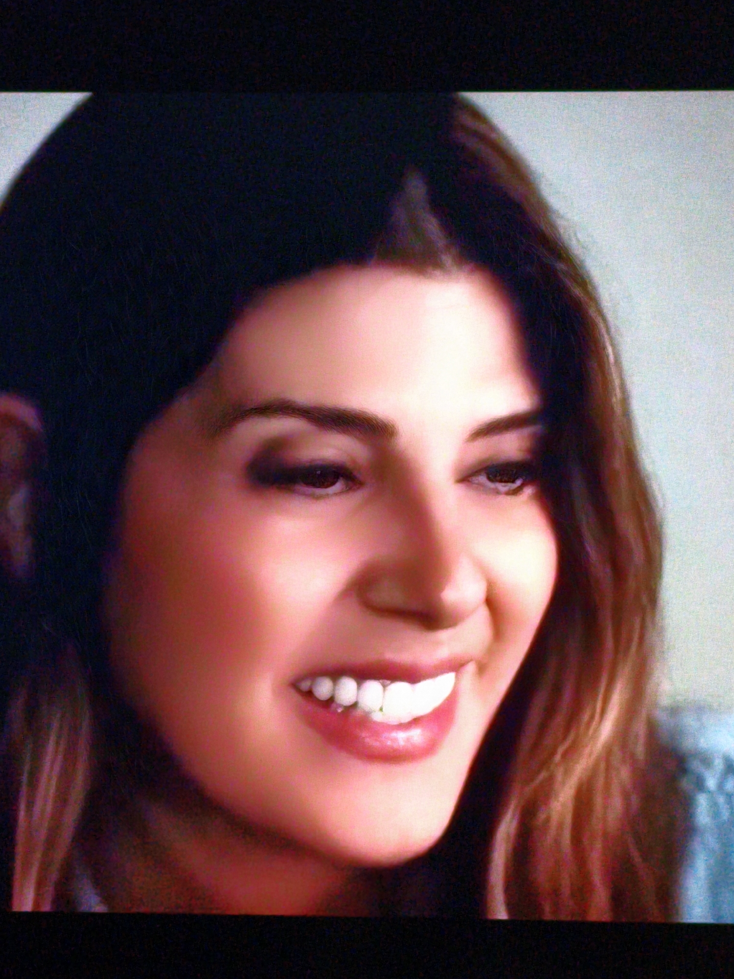 The image is a close-up, slightly grainy, and overexposed shot of a woman's face. She has dark hair that frames her face and fair skin with a rosy hue. Her eyes are dark, and she is wearing dark eyeliner. Her lips are slightly parted, revealing white teeth, and her smile appears to be directed at the camera. The background is a soft, light blue, and there's a hint of a blue garment on her right shoulder. There is no discernible information about the location in Vladivostok, Russia, as the image focuses solely on the woman's face.