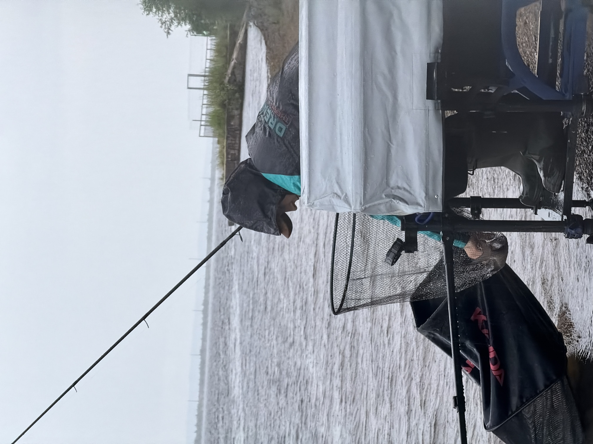 A person is fishing in the Kama River in Chaikovsky, Russia. They are wearing a black rain jacket with a teal accent, a hood, and green rubber boots. The person is holding a fishing rod and a net is nearby, with a fish visible inside it. A black fishing bag is also present, partially submerged in the water. The environment appears overcast and wet, suggesting rain. In the background, there is a shoreline with some vegetation and structures.