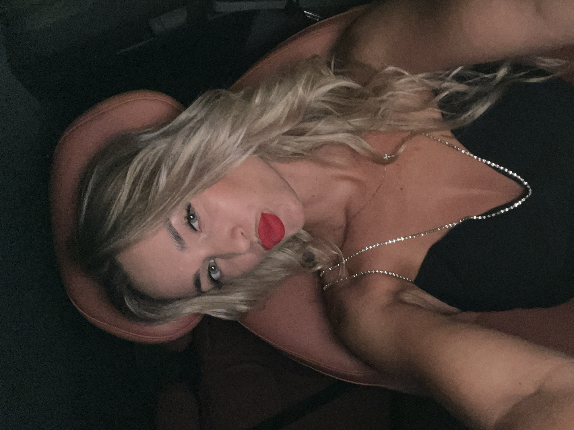 A woman with blonde, wavy hair and red lipstick is taking a selfie in a car. She is wearing a black top with a sparkly necklace. The car interior appears to be luxurious, with leather seats. The lighting suggests it is nighttime. The location of Sharm El Sheikh, Egypt is not evident from the image itself.