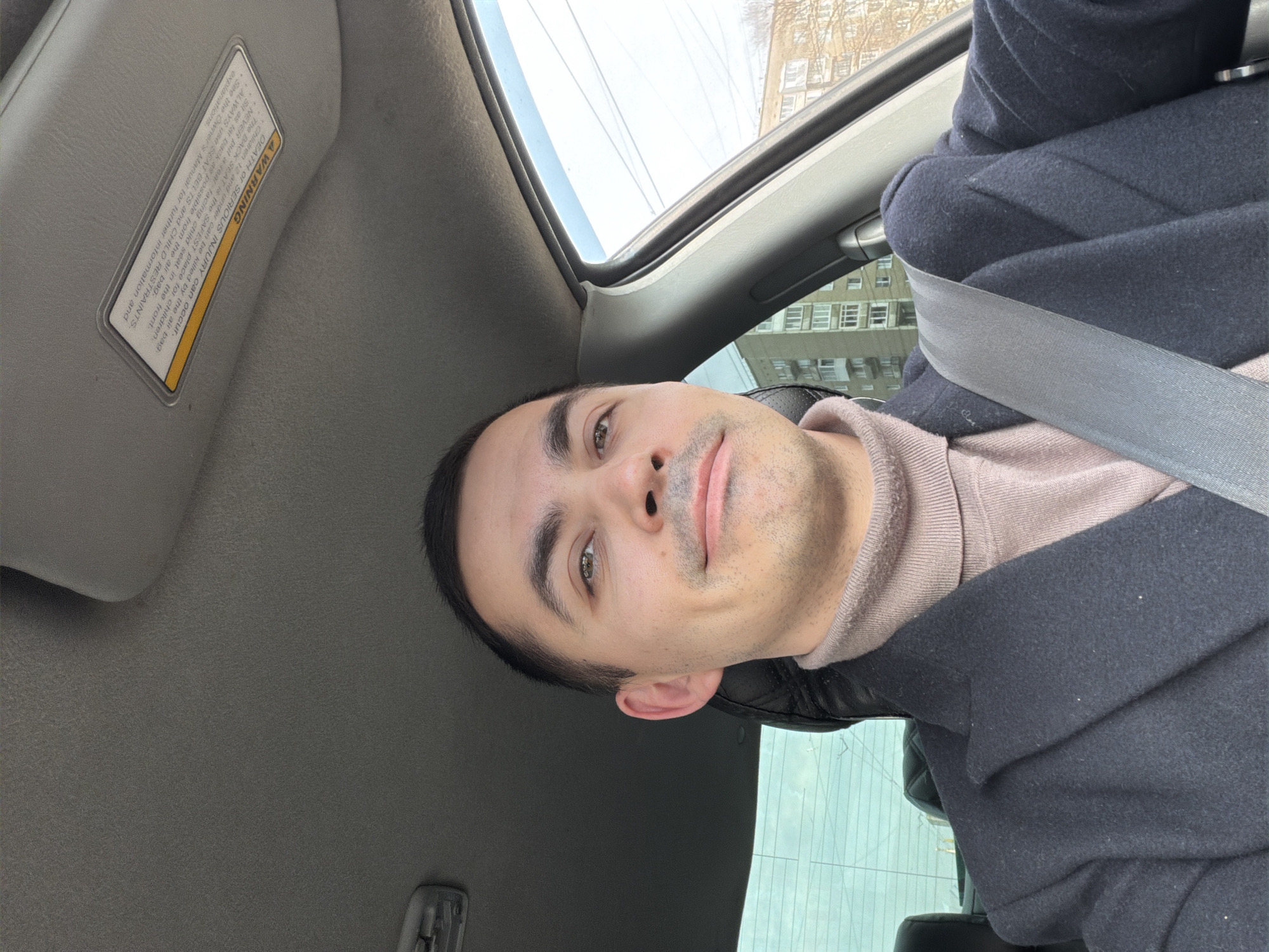 A man wearing a black coat and a seatbelt is inside a car. The car's sun visor is down. Through the car window, a building is visible. The location is Izhevsk, Russia.