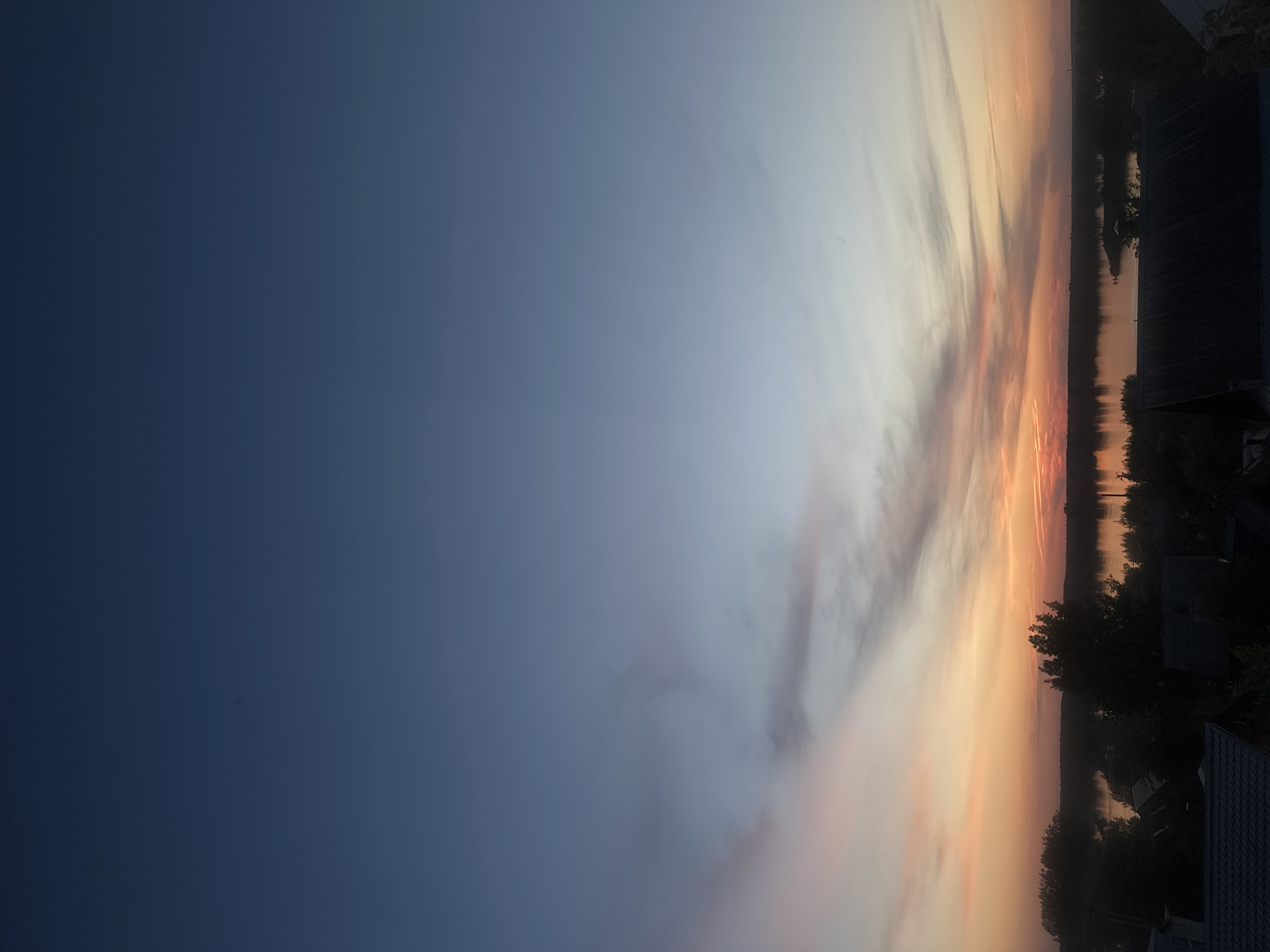 The image depicts a sunset over a body of water. The sky transitions from a dark blue on the left to a light orange and pink on the right, indicating the sun is setting. Clouds are visible in the lighter portion of the sky. Along the horizon, the water reflects the colors of the sunset. The silhouette of trees and possibly rooftops are visible on the right side of the image, indicating a shoreline or built environment. The image orientation is vertical. Based on the prompt, the location is assumed to be in the city of Chaikovsky, Russia.