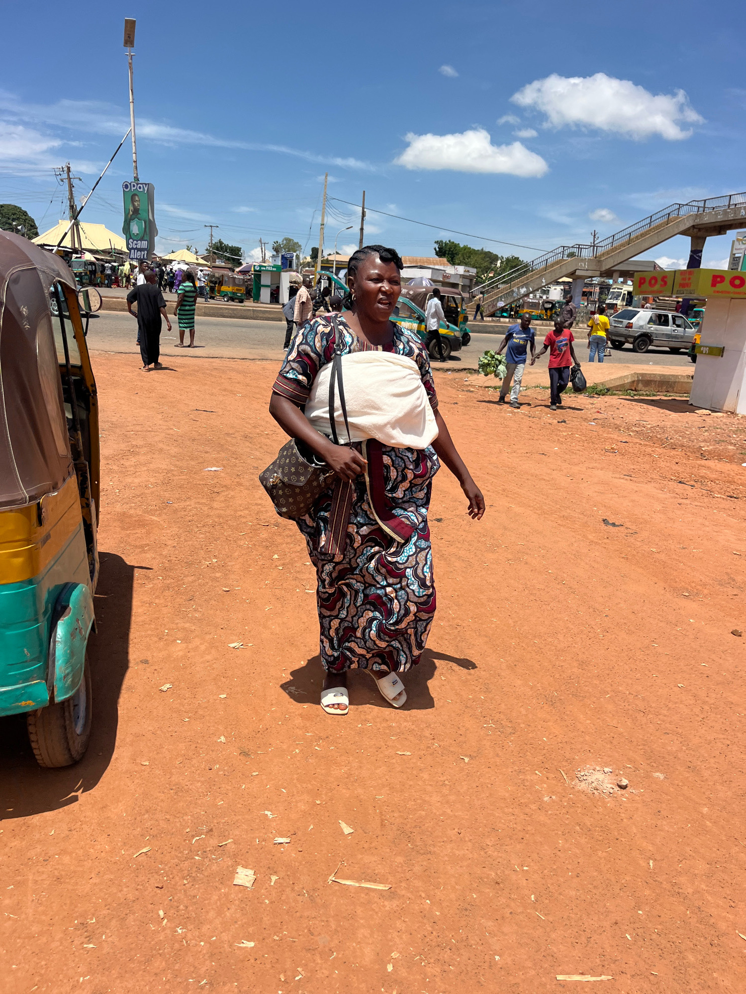 A woman walks across a dirt road in Gura Top, Nigeria, with a baby wrapped in white fabric on her shoulder. She is wearing a vibrantly patterned dress and carrying a designer handbag. In the background, a pedestrian overpass, shops, and various vehicles, including a yellow and green auto-rickshaw, are visible. The sky is blue with scattered clouds.