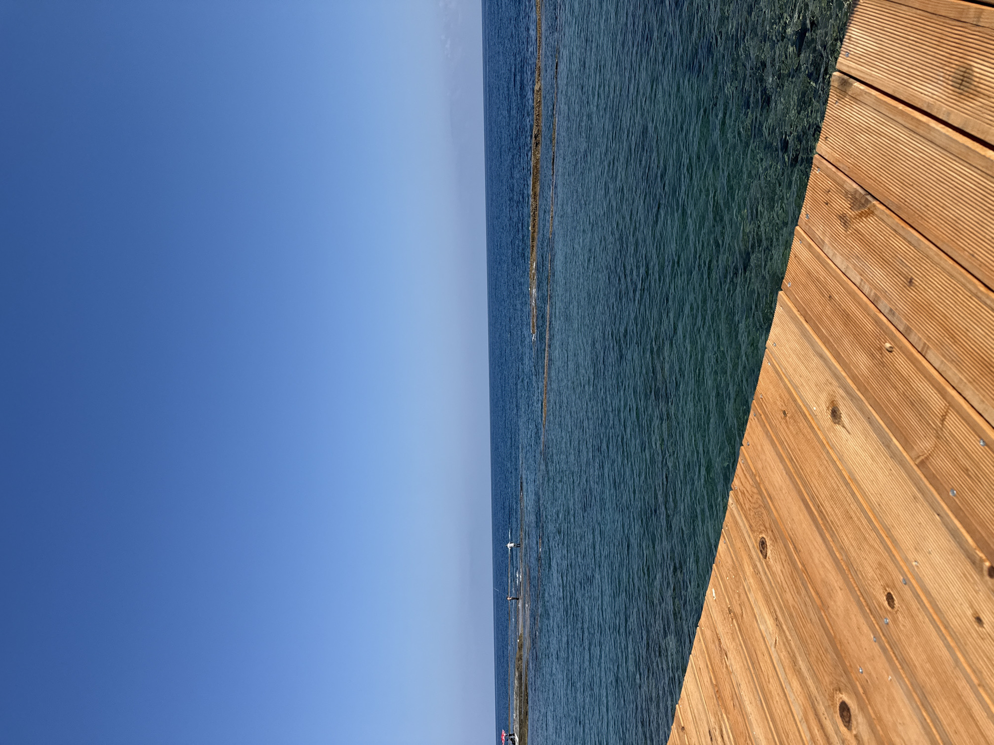 A wooden pier extends into the clear blue water of the sea under a clear blue sky. The pier occupies the lower left portion of the frame. In the distance, a rocky formation breaks the surface of the water, and several people are visible on it. One person is near a red umbrella, and two others appear to be fishing. The water is a vibrant turquoise, with the seabed visible in the shallower areas. The sky is a seamless expanse of blue. The scene suggests a calm and pleasant day at the seaside in Kalogreia, Northern Cyprus.