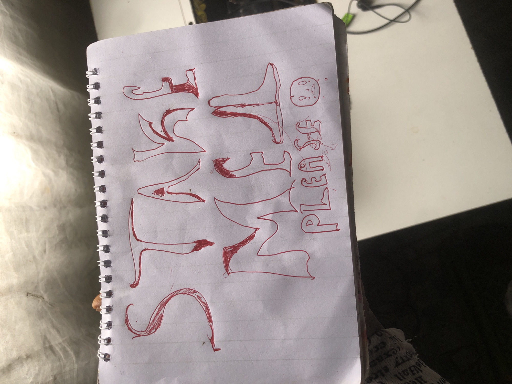 A handwritten note on a spiral-bound notebook displays the words "STAKE ME PLEASE" in red marker. The letters are large and stylized, with some appearing to have decorative elements. Below the text, a simple drawing of a distressed face with wide eyes and a downturned mouth is also sketched in red. The notebook page is lined and white. The image is taken from a high angle, showing the notebook lying on a surface. The background is out of focus but appears to be indoors with some light sources. The notebook is positioned on a desk or table, with a portion of a black object and a white surface visible. There is no discernible location in Ado, Nigeria visible in the image.