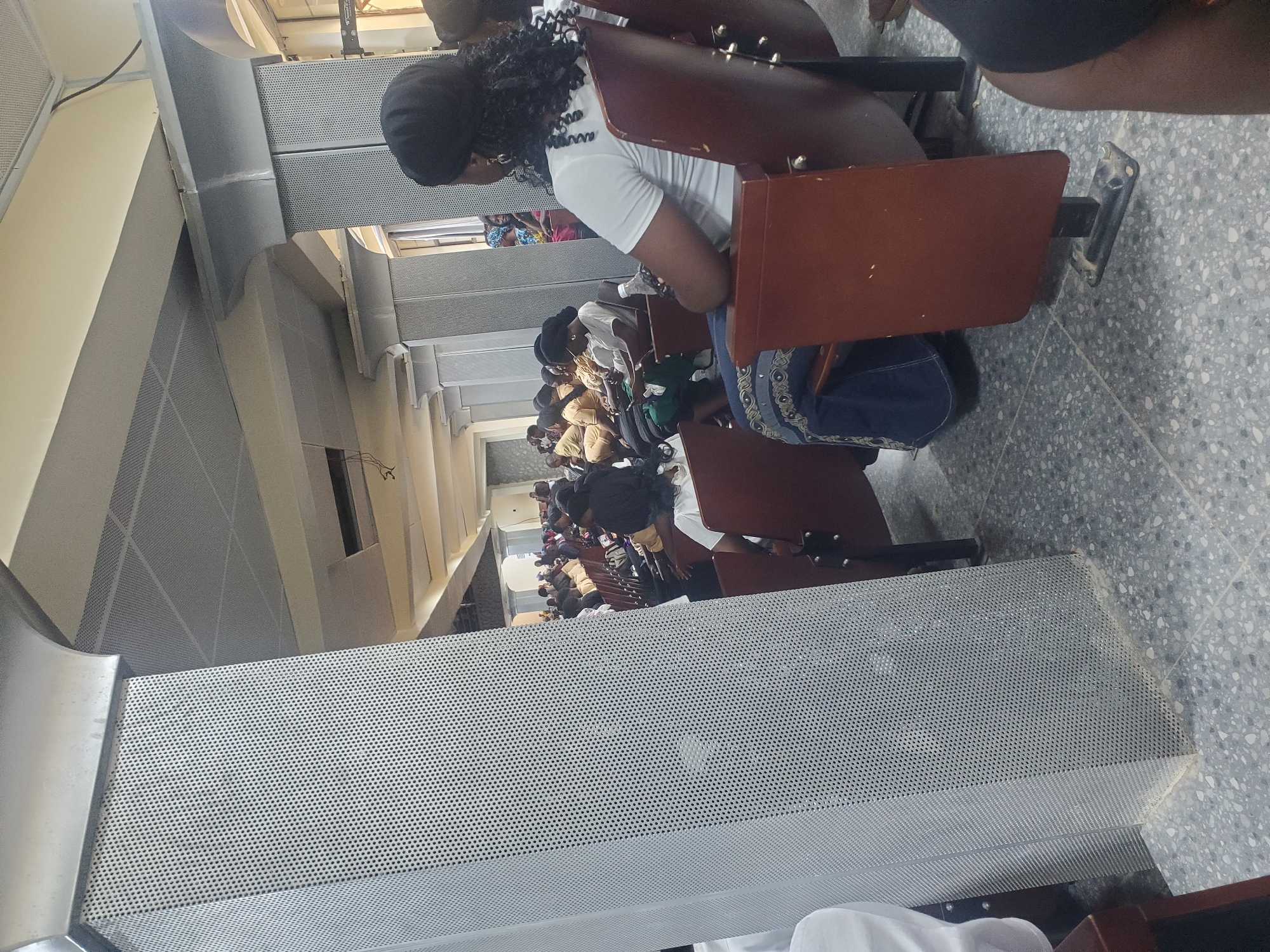In Kason, Nigeria, a dimly lit indoor space, possibly a church or conference hall, accommodates a gathering of people seated in rows of brown chairs. The hall features a perforated metal pillar in the foreground, a suspended acoustic tile ceiling, and decorative architectural elements. A woman wearing a white top and dark headscarf is seated in the front row, facing away from the camera. Other individuals, mostly women dressed in modest attire, are visible throughout the hall, some looking ahead, others engaged with their phones or conversing. The overall atmosphere appears calm and attentive.