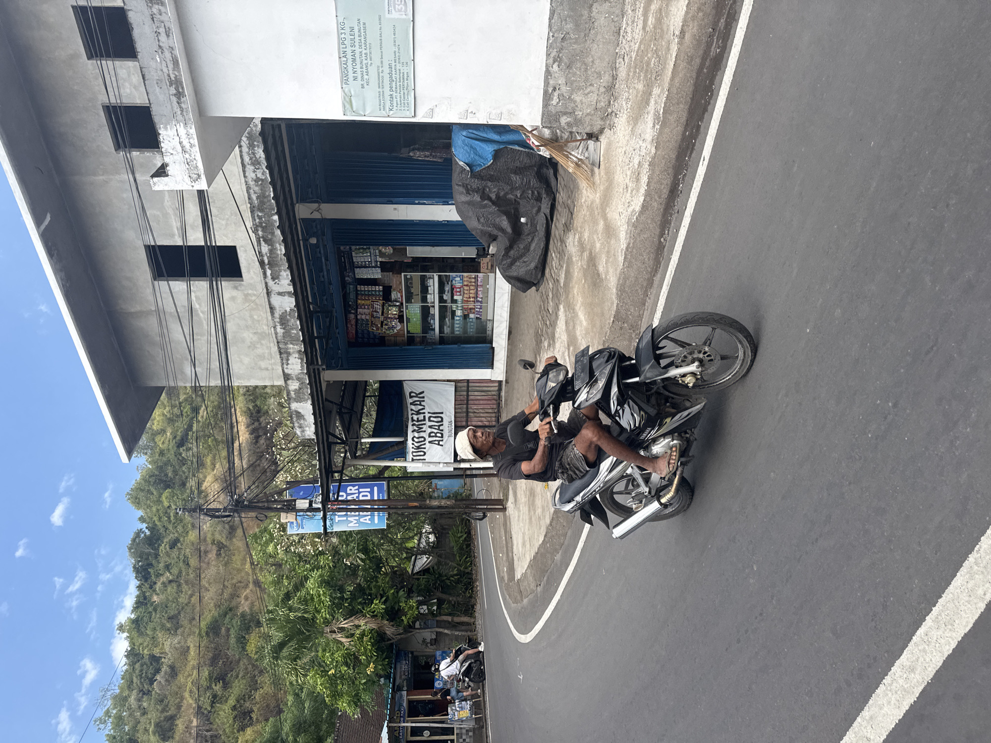 An elderly man wearing a white head covering, a dark t-shirt, and shorts rides a black motorcycle down a paved road. The man is facing left and looking forward, his right foot is on the right footrest, and his left foot is resting on the ground near the rear wheel. He has a thin build and appears to be of Asian descent. The road curves to the left. On the right side of the road is a building with a gray facade, a white awning, and a blue shuttered storefront. A large black tarp covers an object in front of the building. A sign above the tarp reads "PANGKALAN LPG 3 KG - NI NYOMAN SULENI." To the left of the road, there is a smaller building with an open blue shutter and several shelves stocked with goods. A blue sign above this store reads "TUK ME AR AB ADI". Further down the road, a few people are standing near a building with a sign that says "ANJ EAST OF TAXI". A green, treed hillside rises behind the buildings. The sky is blue with scattered white clouds.