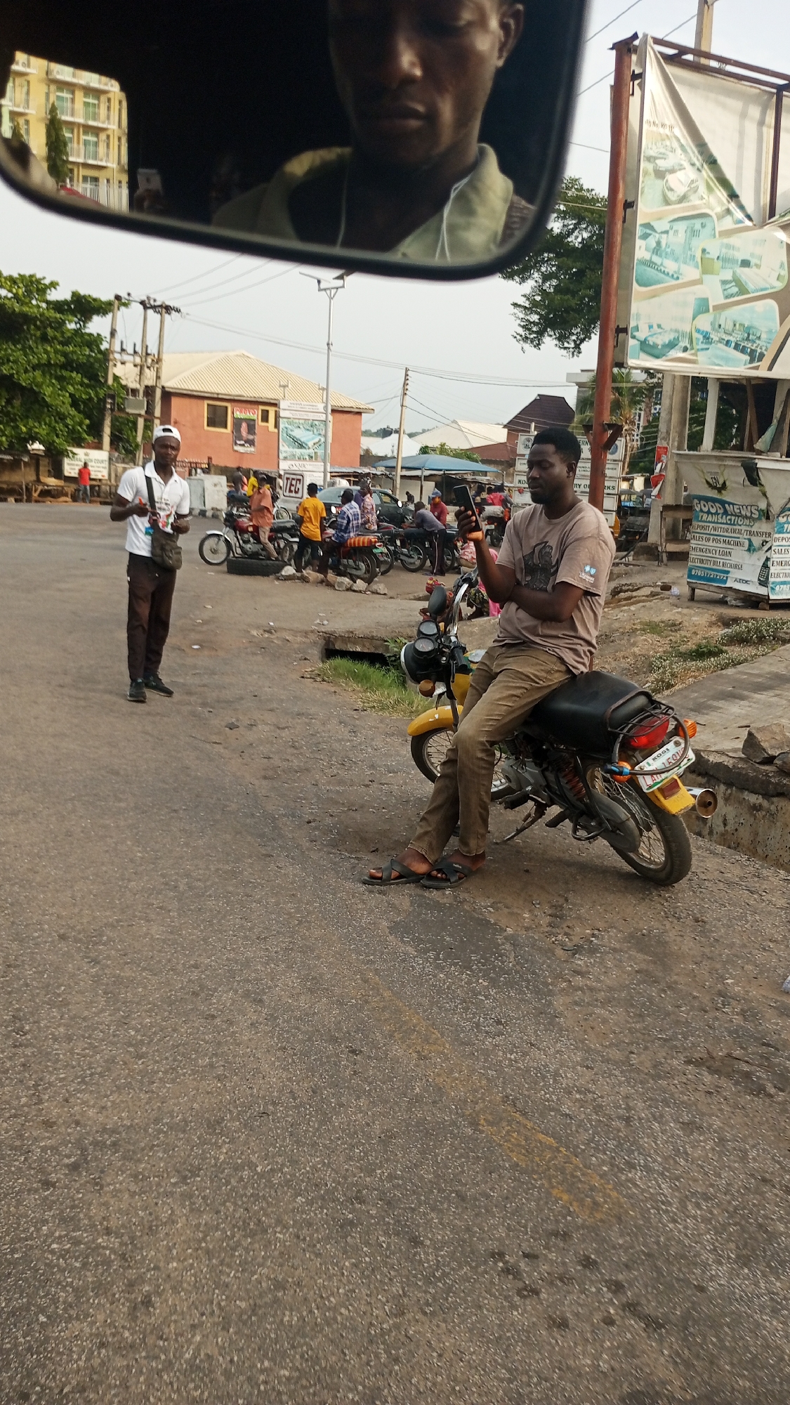 This image captures a street scene in Zango-ndaji, Nigeria, under bright daylight. The primary subject in the foreground is the reflection of a man's face, likely the driver or photographer, visible in a rearview mirror. His expression is neutral as he looks downwards.
In the midground, a man sits sideways on a yellow and black motorcycle parked by the roadside, engrossed in his smartphone. He wears a light brown t-shirt, brown trousers, and dark sandals. The motorcycle's license plate is partially visible, showing "DE KOGI". To his left, another man stands on the asphalt road, wearing a white cap, white t-shirt, and brown trousers, holding a small white object, possibly a phone, in his hand while a bag hangs over his shoulder.
The background reveals a bustling street environment. Multiple motorcycles are parked or in motion, with several people gathered around them, suggesting a common waiting or meeting point. Various buildings with different architectural styles and roof colors line the street. Utility poles and power lines crisscross the sky.
On the right side of the frame, a large signboard features graphics and text. Visible text includes "GOOD NEWS TRANSACTIONS", listing services such as "DEPOSIT/WITHDRAWAL/TRANSFER", "SALES OF POS MACHINE", "EMERGENCY LOAN", and "ELECTRICITY BILL RECHARGE", along with a phone number "07051721294 ADC". Another smaller sign shows "TEC". The road surface is paved but appears worn, with a faint yellow line near the bottom right. The overall atmosphere is that of a typical, busy urban or semi-urban area.