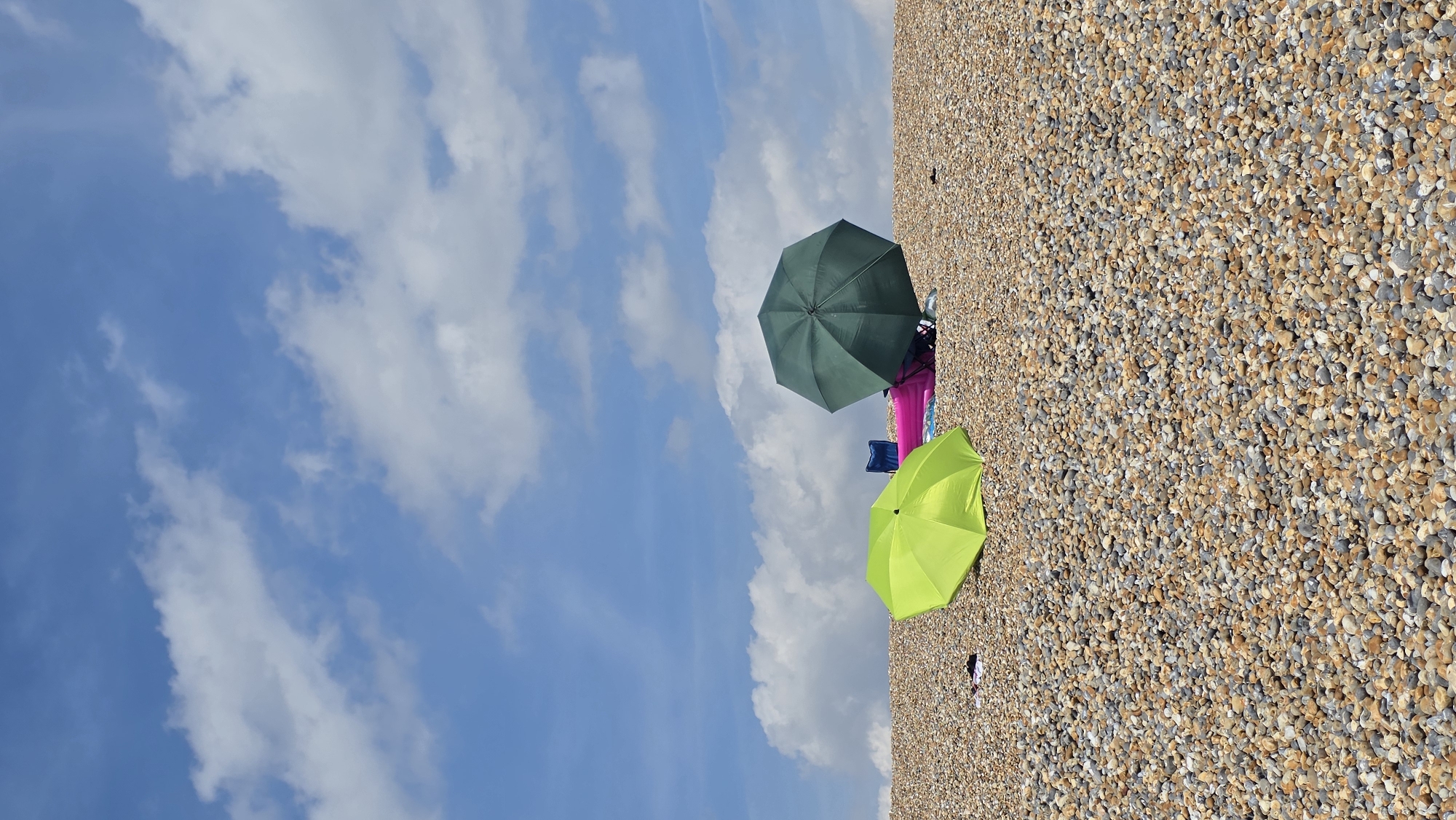 On a bright, sunny day, two beach umbrellas are set up on a pebble beach. One umbrella, bright lime green, is positioned in the foreground, while a darker green umbrella stands further back, partially obscuring some beach accessories. Beneath the darker umbrella, a bright pink inflatable mattress and a blue folding chair are visible, suggesting a relaxing day at the beach. The sky above is a vivid blue, dotted with fluffy white clouds, indicative of fair weather. The beach itself is covered in a dense layer of smooth, multi-colored pebbles. This scene is located on the St Leonards coastline in the United Kingdom.