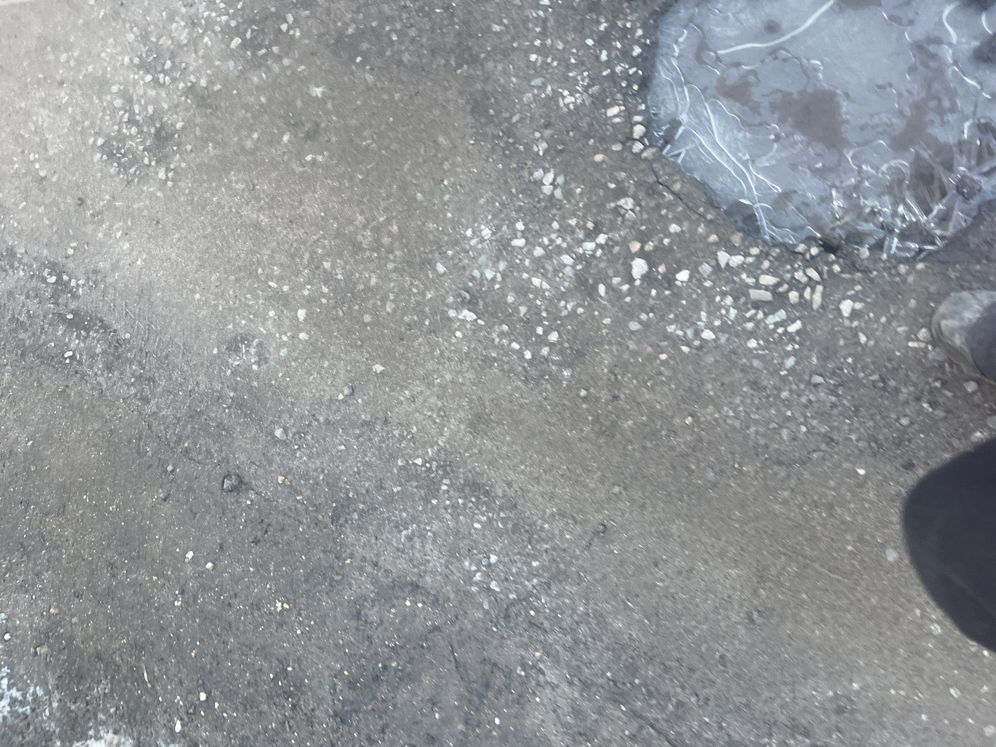 The image presents a top-down view of a grey, textured ground surface identified as being in the city of Podolsk, Russia. The surface appears to be weathered asphalt or concrete, characterized by numerous small cracks, scattered light-colored gravel, and darker debris. In the bottom right quadrant, a shallow, irregular depression contains a body of water that is partially covered by a thin, transparent layer of ice. The ice exhibits distinct, sinuous formations and fractured edges. A portion of a dark-colored shoe or boot, specifically the toe and front instep of what appears to be a left foot, is visible in the bottom left corner of the frame. A dark, diffuse shadow, likely cast by the photographer, extends from the bottom center towards the middle of the image, partially obscuring the visible shoe. No other individuals or specific objects are discernible.