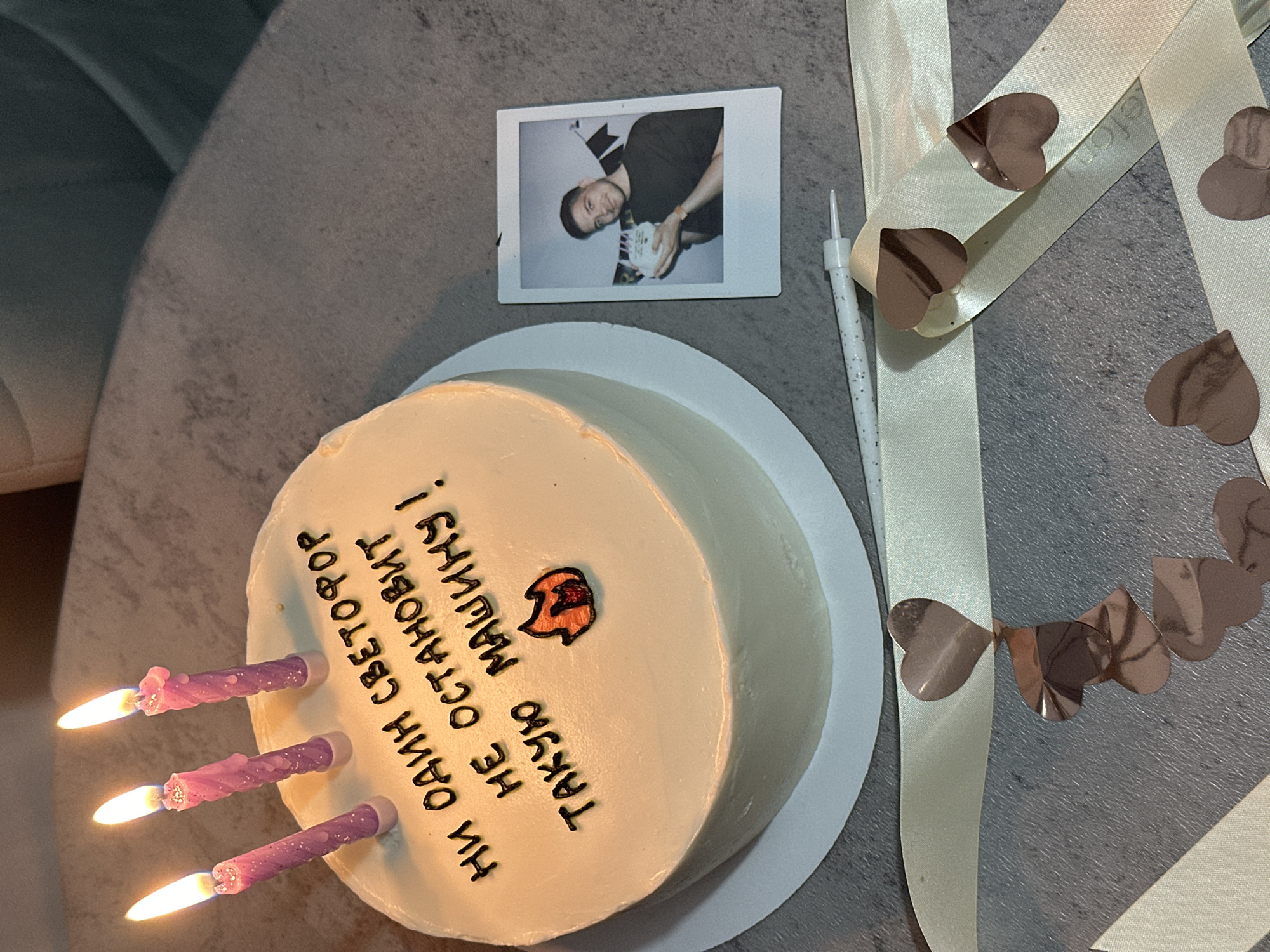 The image captures a birthday celebration, possibly in Kazan, Russia, as the cake is inscribed with Russian text. The scene unfolds on a grey tabletop, featuring a round cake adorned with light-colored frosting, black lettering, and a small orange decoration. Three lit purple candles are placed on the left side of the cake. A white plate supports the cake. Adjacent to the cake, there is a Polaroid photograph of a man holding a similarly frosted cake. A white candle and a cream-colored ribbon decorated with small heart-shaped embellishments are placed next to the cake. The surrounding environment is blurred, suggesting a domestic setting.