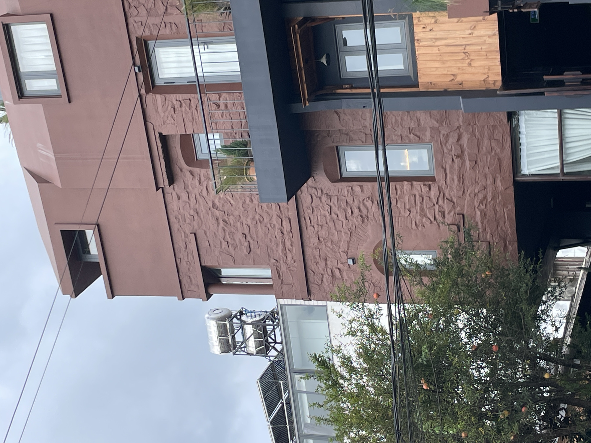 The image shows the exterior of a multi-story building with a dark red, textured facade, possibly made of stone or brick. The building has several windows with grey frames and white curtains, and some balconies with metal railings. There is a dark grey overhang on the lower part of the building, supported by dark pillars, with a wooden panel underneath and a hanging lamp. To the left of the main building, a portion of a white building with large windows is visible, and a tree with several red fruits, likely pomegranates, is in the foreground. A solar water heater and solar panels are on the roof of the white building. Overhead, several black wires are strung across the frame, and the sky is overcast. The architectural style suggests a modern design with rustic elements. The scene appears to be located in Lâm Viên - Đà Lạt, Vietnam, given the presence of pomegranate trees and the common use of solar water heaters in the region.