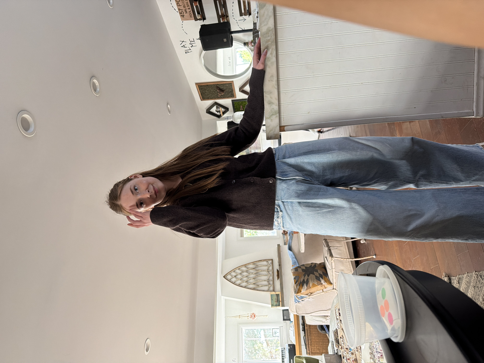 The image shows a young woman indoors in what appears to be a home. She is wearing a dark-colored cardigan and light blue jeans. The interior is well-lit with white walls and what seems to be recessed lighting. Decorative items and furniture, including a sofa, are visible in the background, suggesting a living space. Based on the handwritten "457 PC MALIBU CO. POOL & SPA SET" on the wall, the location is likely Malibu, California, United States. A table with plastic containers is in the foreground. The camera angle is from above.