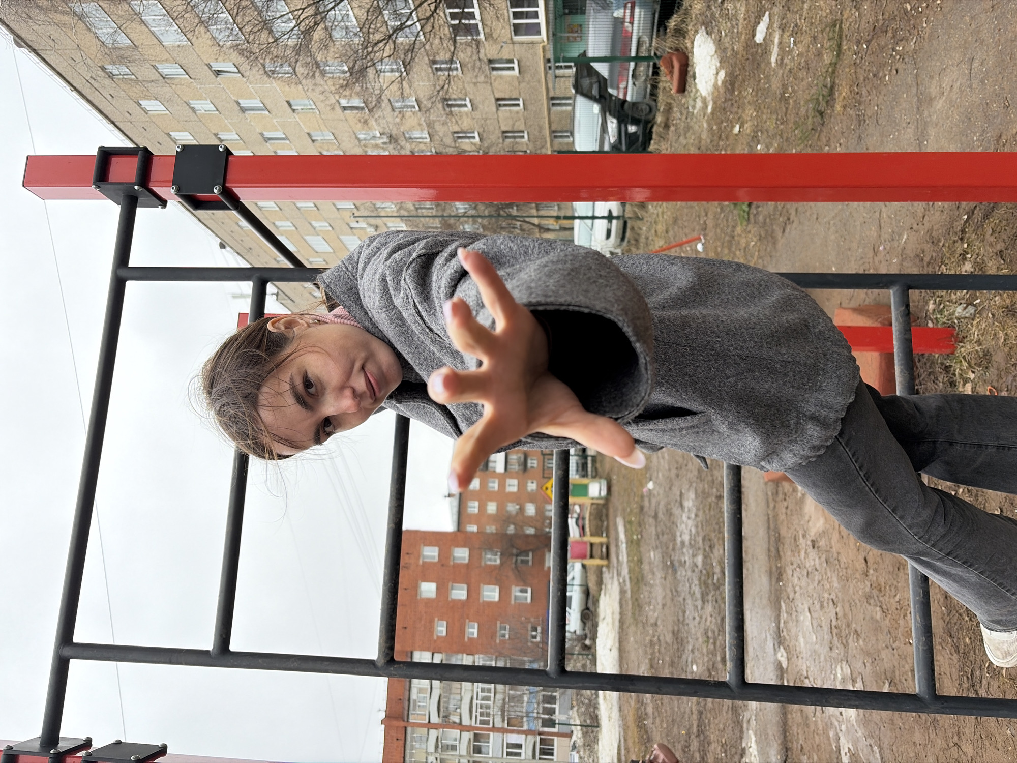 A woman is seen climbing on a metal playground structure outdoors. The structure is painted black and red. She is wearing a gray coat and gray jeans with white sneakers. Buildings are visible in the background, consistent with an urban environment. The location is Izhevsk, Russia. The playground is located between two buildings, and there is dirt and sparse vegetation on the ground. The sky is overcast. The woman is reaching out her hand towards the camera.