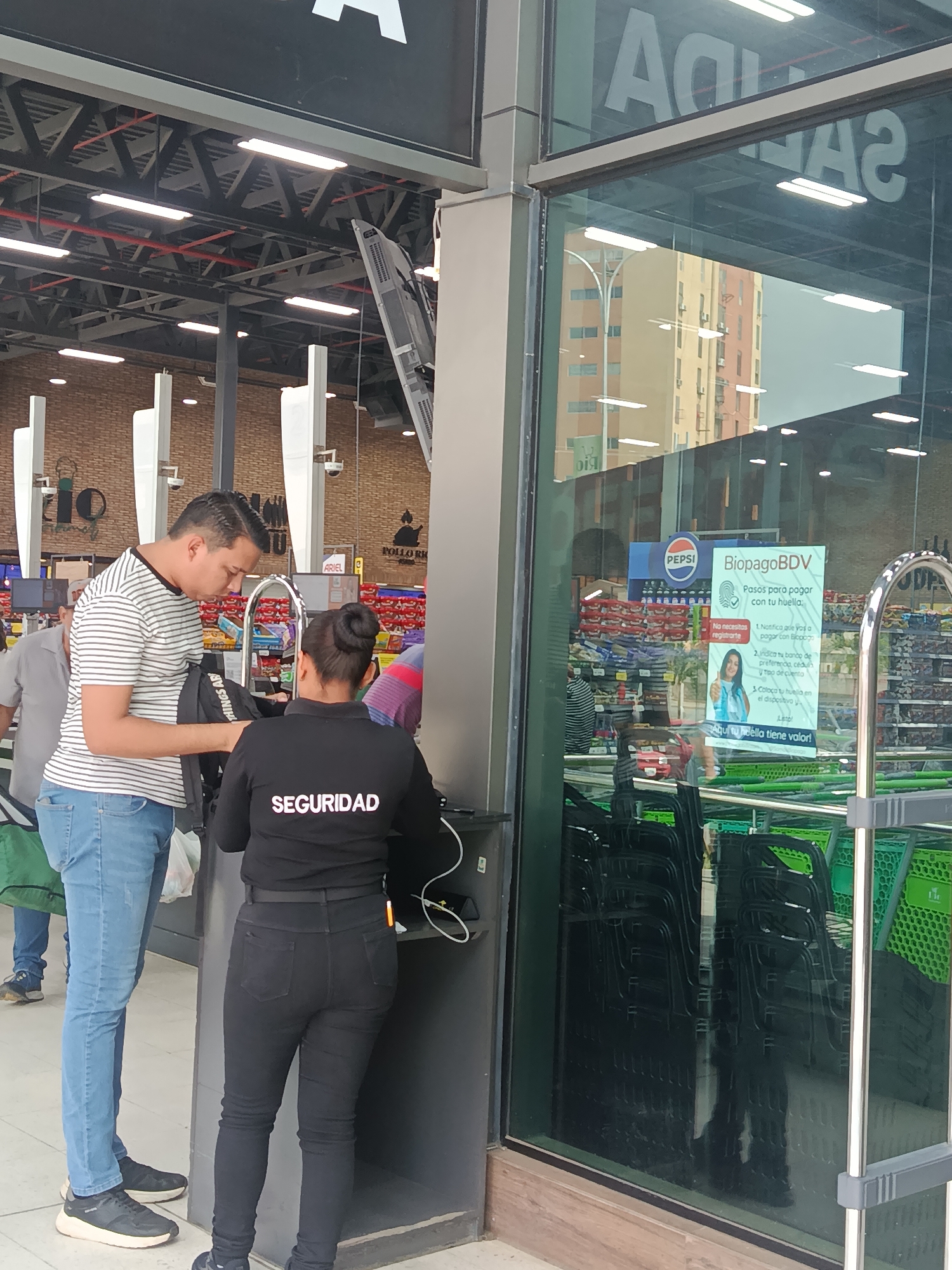 This image captures a bustling scene at the entrance or exit of a supermarket in Juan Griego, Venezuela, during what appears to be daytime.
In the foreground, a man in a striped t-shirt, blue jeans, and dark sneakers is interacting with a security checkpoint or service counter. He is looking down, seemingly placing or retrieving an item from a black bag that bears the partial text "THINGS AR." To his right, a female security guard is seen from behind, wearing a black uniform shirt with "SEGURIDAD" printed prominently in white across her back, black trousers, and dark sneakers. She is standing at the counter, possibly attending to the man or operating a device.
The background reveals the interior of a large retail space with an industrial aesthetic, featuring exposed metal trusses, bright fluorescent lighting, and red piping on the ceiling. Various supermarket shelves and displays are visible, with signs for "POLLORA" (likely a food item like chicken) and "ARIEL" (a brand of detergent). Other patrons are partially visible in the background, including a man in a gray t-shirt to the left.
On the right side of the image, a large glass partition reflects the exterior surroundings, including a multi-story building with beige and orange facades, suggesting an urban setting. Reversed text, possibly "SALIDA" (Exit), is also reflected on the glass. Attached to the glass from inside is a poster advertising "BiopagoBDV," a biometric payment system. The poster, featuring a "PEPSI" logo at the top and a smiling woman, explains the "Pasos para pagar con tu huella" (Steps to pay with your fingerprint), noting "No necesites registrarte" (No need to register). The steps include notifying the cashier, indicating bank details and ID ("cedun" likely meaning "cédula"), and placing a fingerprint on the device. The slogan "Aqui tu hdella tiene valor!" (Here your fingerprint has value!) is also visible. Through the glass, stacks of black and green shopping carts are visible.