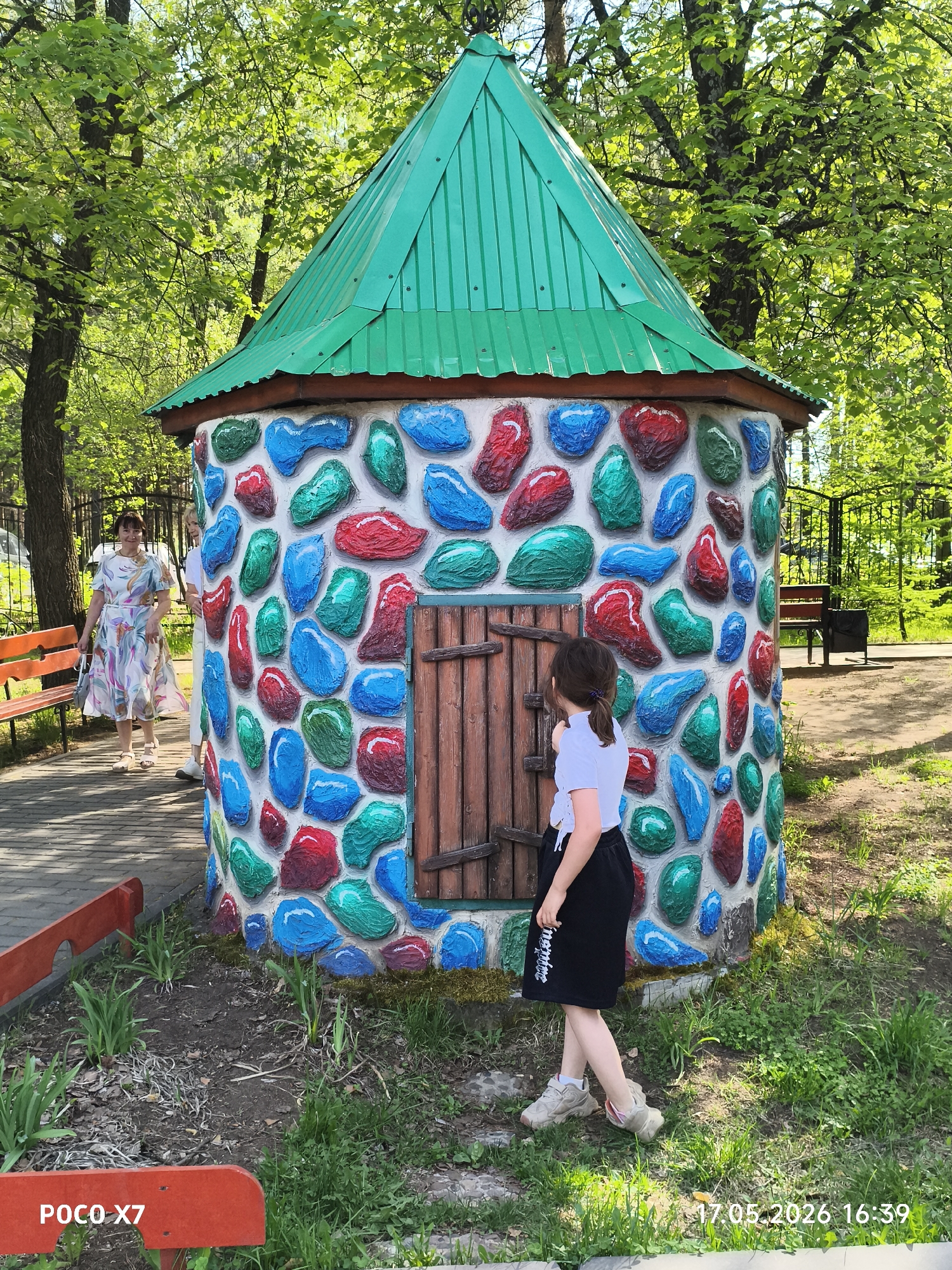The image shows a young girl standing in front of a small, colorful, stone-like structure with a green roof. The structure appears to be a playhouse or a decorative feature in a park or public space. The walls are adorned with painted "stones" in shades of red, blue, and green. The structure has a small wooden door.
In the background, a woman in a floral dress walks along a paved path near a bench. Trees surround the scene. The photograph was taken in Chaikovsky, Russia. The bottom right corner of the image shows a timestamp of 17.05.2026 16:39 and the bottom left corner says “POCO X7”.