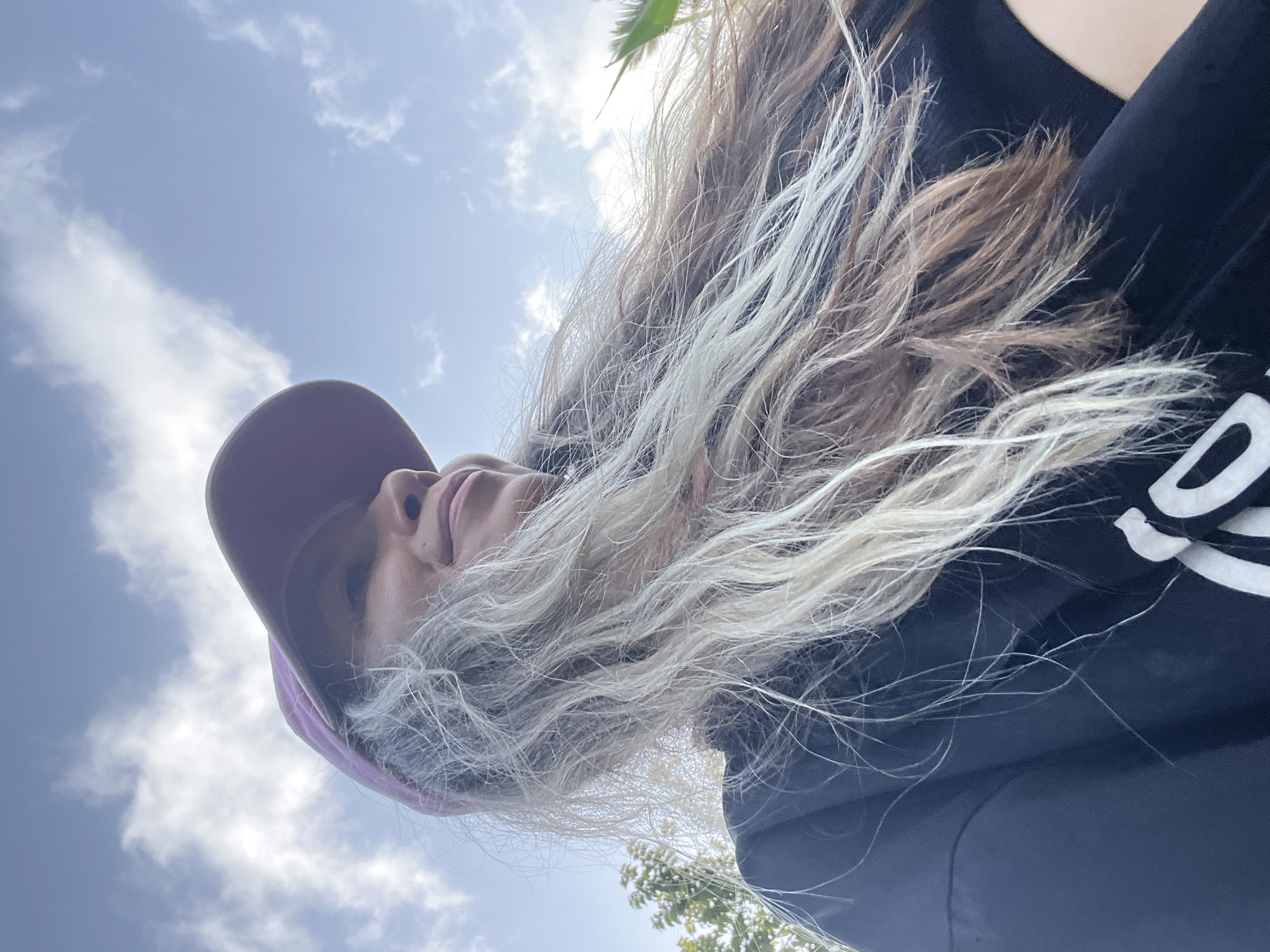 A low-angle, outdoor, close-up selfie captures a woman wearing a purple baseball cap and a black t-shirt. Her long, blonde and brown hair is blown to the side by the wind, obscuring part of her face. She is smiling and looking off to the right, with a bright blue sky and scattered white clouds in the background. The image appears to have been taken during the day, as indicated by the natural lighting. The background also shows hints of greenery, suggesting a natural or park-like setting.